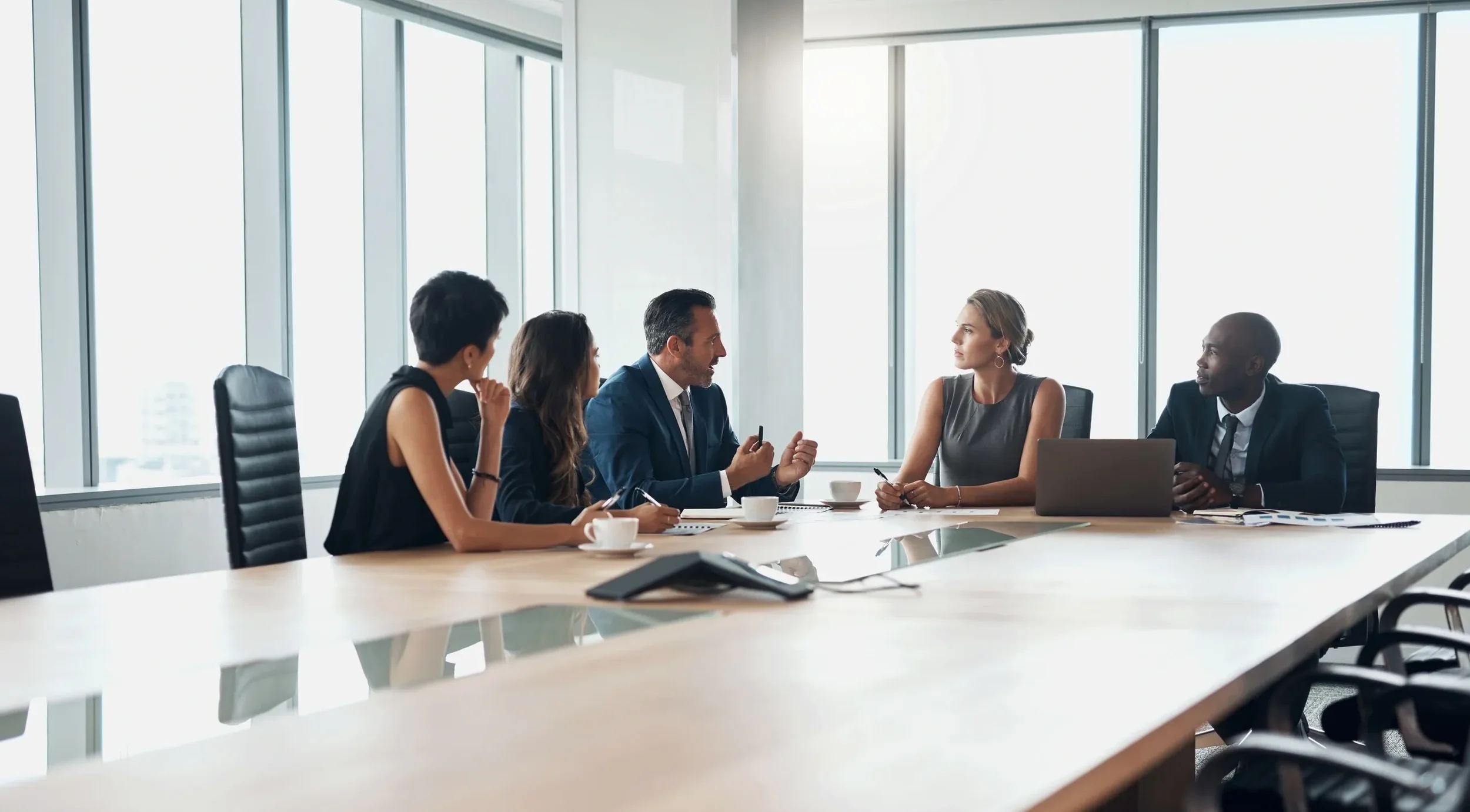 Six people sitting around a conference table in a modern office with large windows, engaged in a discussion.