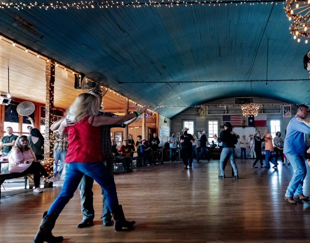 People dancing in Twin Sisters Dance Hall decorated with string lights and flags, with some sitting and watching.