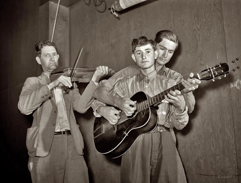 Three men playing musical instruments: one with a violin, two with a guitar, standing against a wooden wall.
