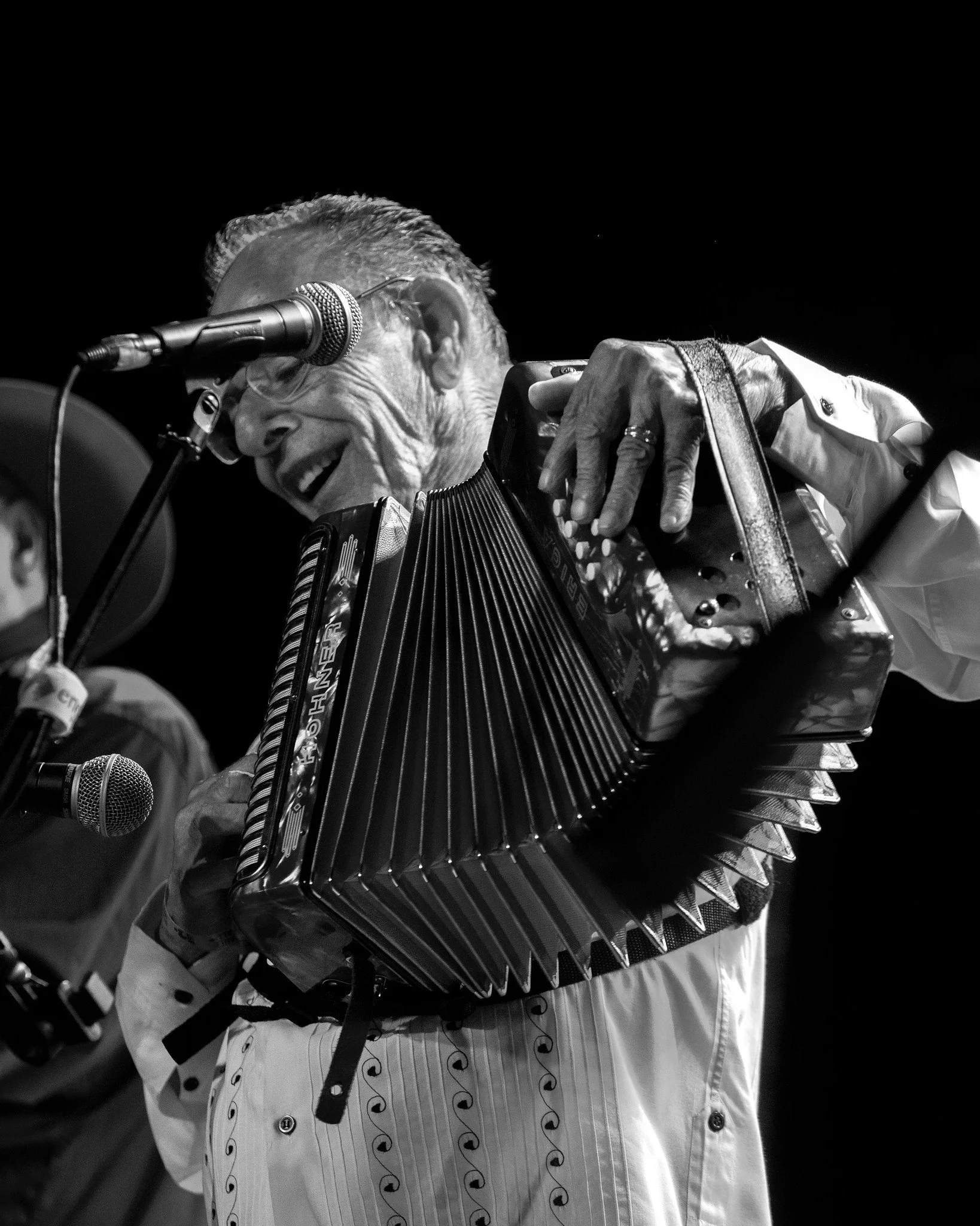 SantiagoJimenez, Jr. playing an accordion on stage, smiling and wearing a patterned shirt, with a microphone nearby and a dark background.