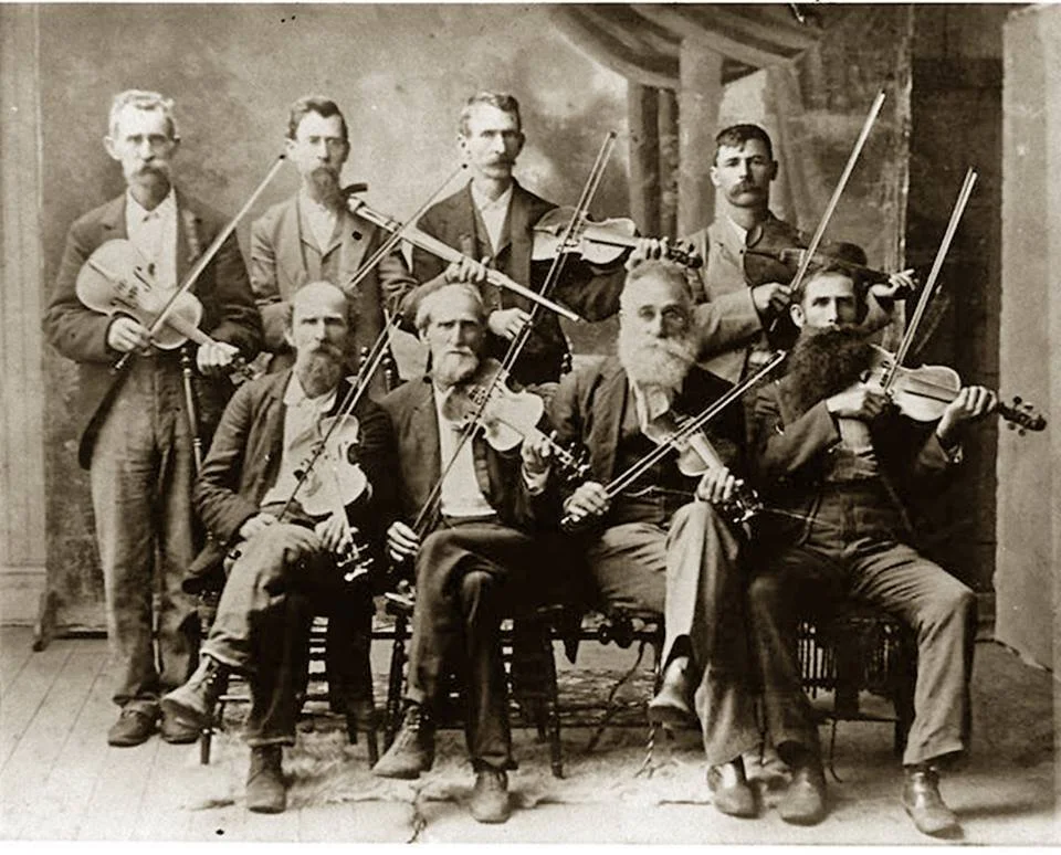 Black and white photo of nine men in old-fashioned clothing, holding and playing violins and a viola, posed in a room with wooden floor and backdrop.