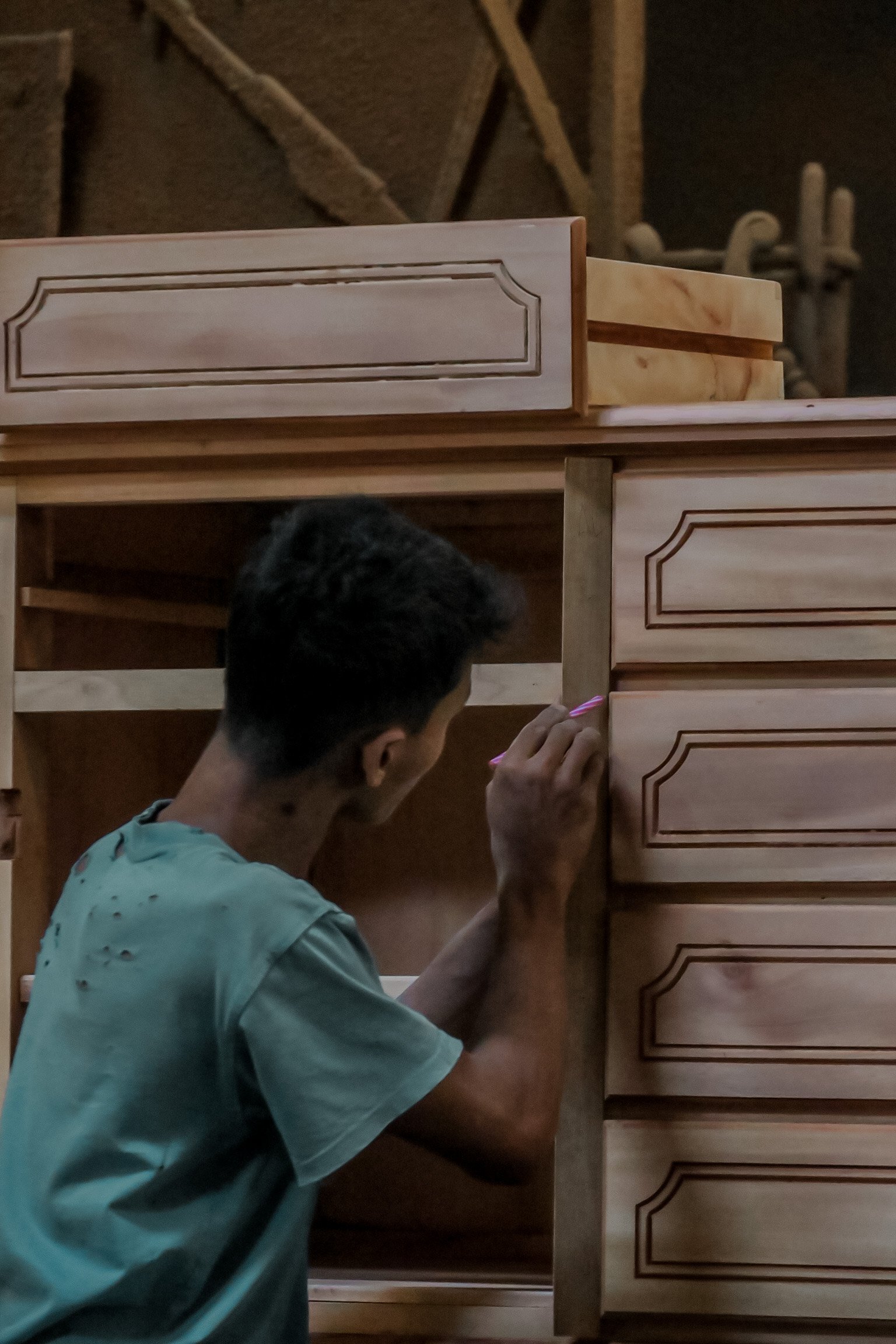 A person with short dark hair painting a wooden cabinet with a small brush in a workshop. Teak Furniture Jepara, wholesale Indonesian teak furniture, custom outdoor teak furniture manufacturer, Jepara teak furniture supplier. 