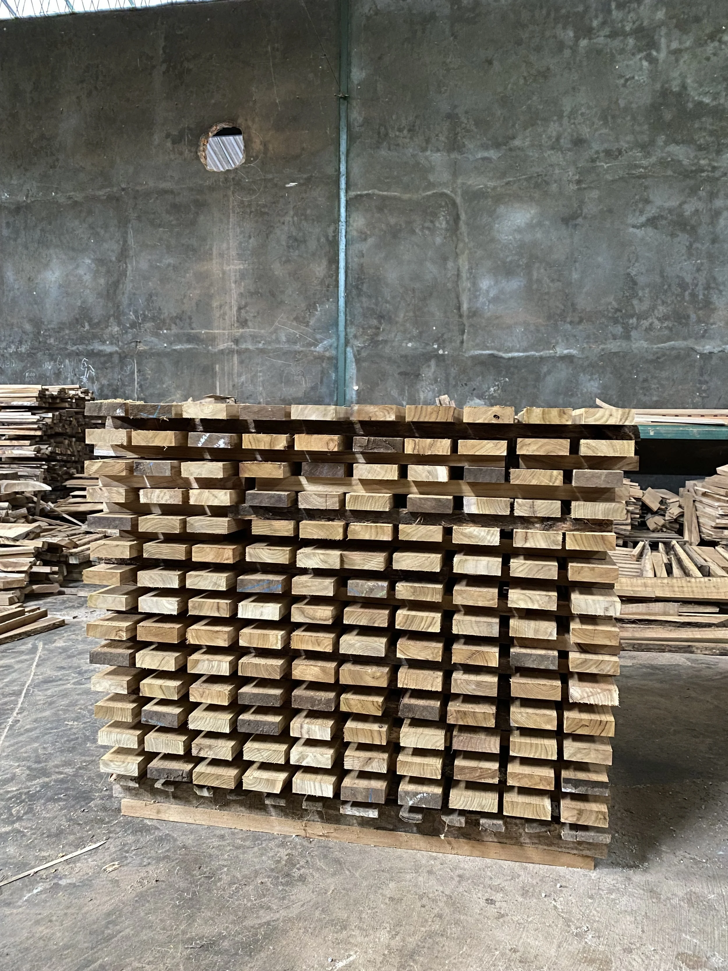 Stacked wooden planks in a warehouse or workshop with a plain concrete wall in the background.