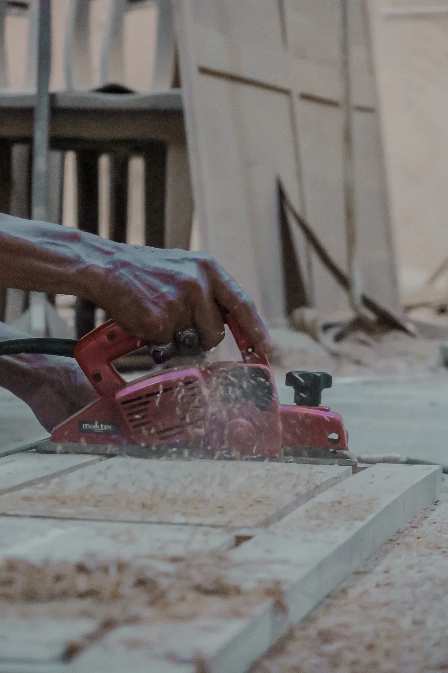 Close-up of a person's hand holding a red power saw, cutting a large sheet of material, with a background chairs and a wall. Teak Furniture Jepara, wholesale Indonesian teak furniture, custom outdoor teak furniture manufacturer, Jepara teak furniture