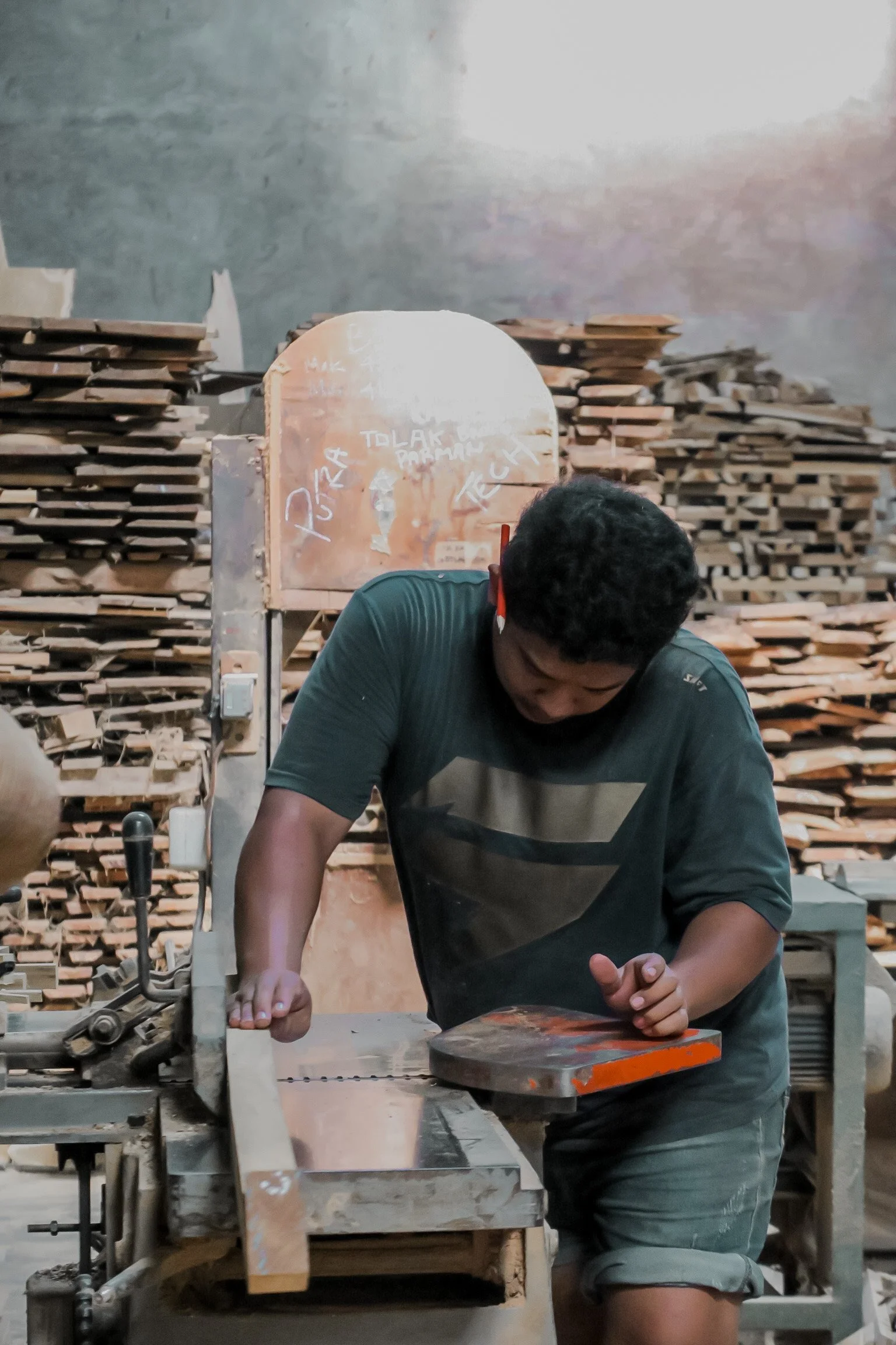 A person working on a woodworking project in a workshop surrounded by stacks of wood and tools. Teak Furniture Jepara, wholesale Indonesian teak furniture, custom outdoor teak furniture manufacturer, Jepara teak furniture supplier. 