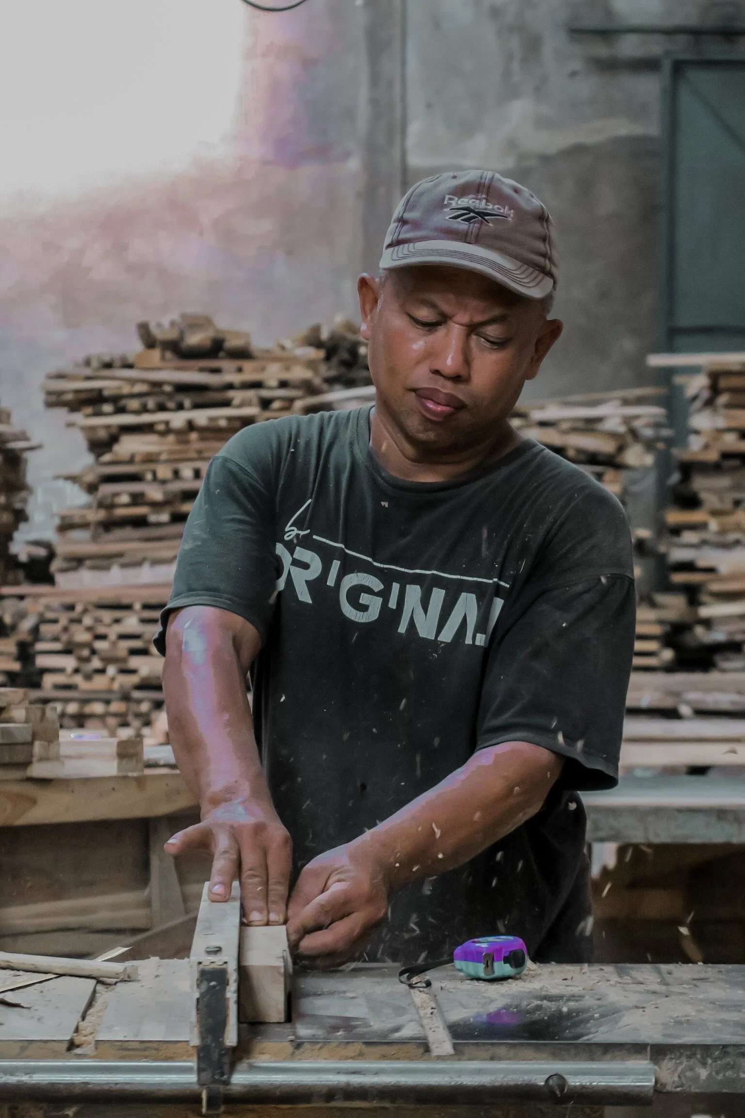 A man wearing a Reebok cap and a black t-shirt working on a woodworking project in a workshop, using a hand saw on a piece of wood with stacks of wooden planks in the background.Teak Furniture Jepara, wholesale Indonesian teak furniture, custom outdo