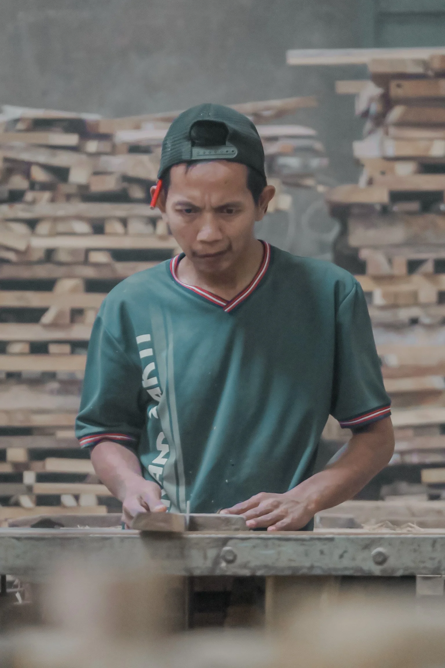 Piles of teak wood planks are stacked in the background, and dust or smoke is visible in the air. Teak Furniture Jepara, wholesale Indonesian teak furniture, custom outdoor teak furniture manufacturer, Jepara teak furniture supplier. 