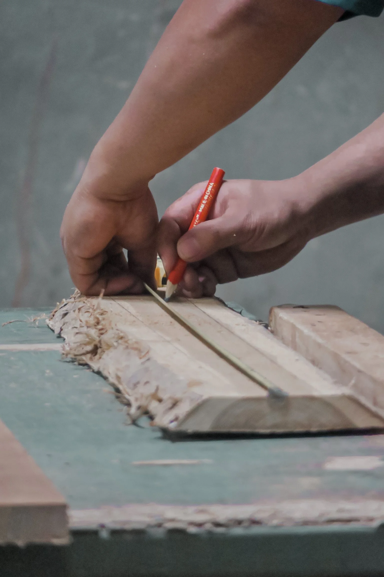 Close-up of a person using a red marker to draw or write on a piece of wood, with wood shavings visible on a green surface. Teak Furniture Jepara, wholesale Indonesian teak furniture, custom outdoor teak furniture manufacturer, Jepara teak furniture 
