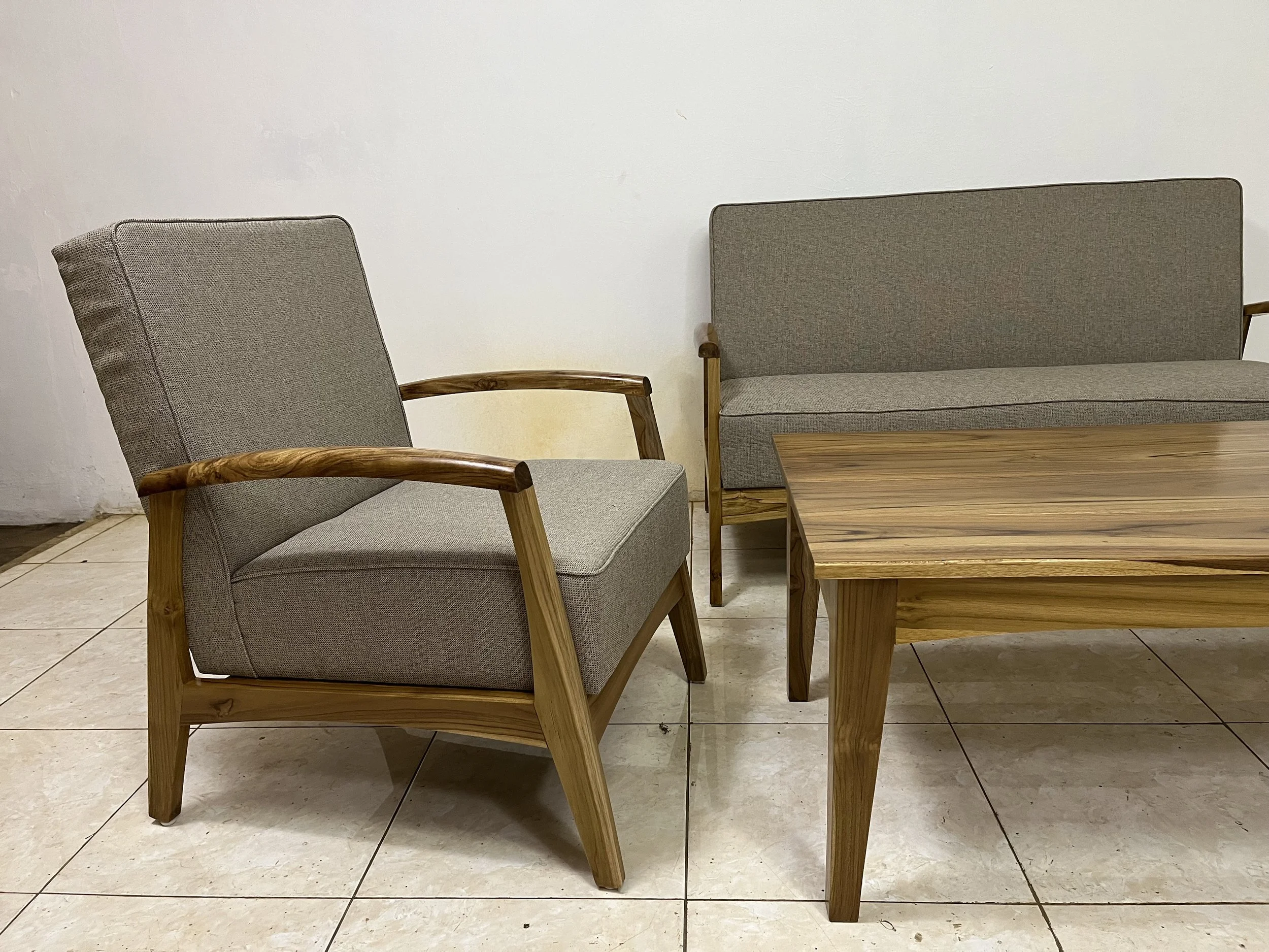 A room with two chairs and a wooden table. One chair has a wooden frame with gray fabric upholstery, and the other is a gray upholstered bench with wooden armrests. The table is rectangular and made of wood. The floor is tiled, and the wall is plain 