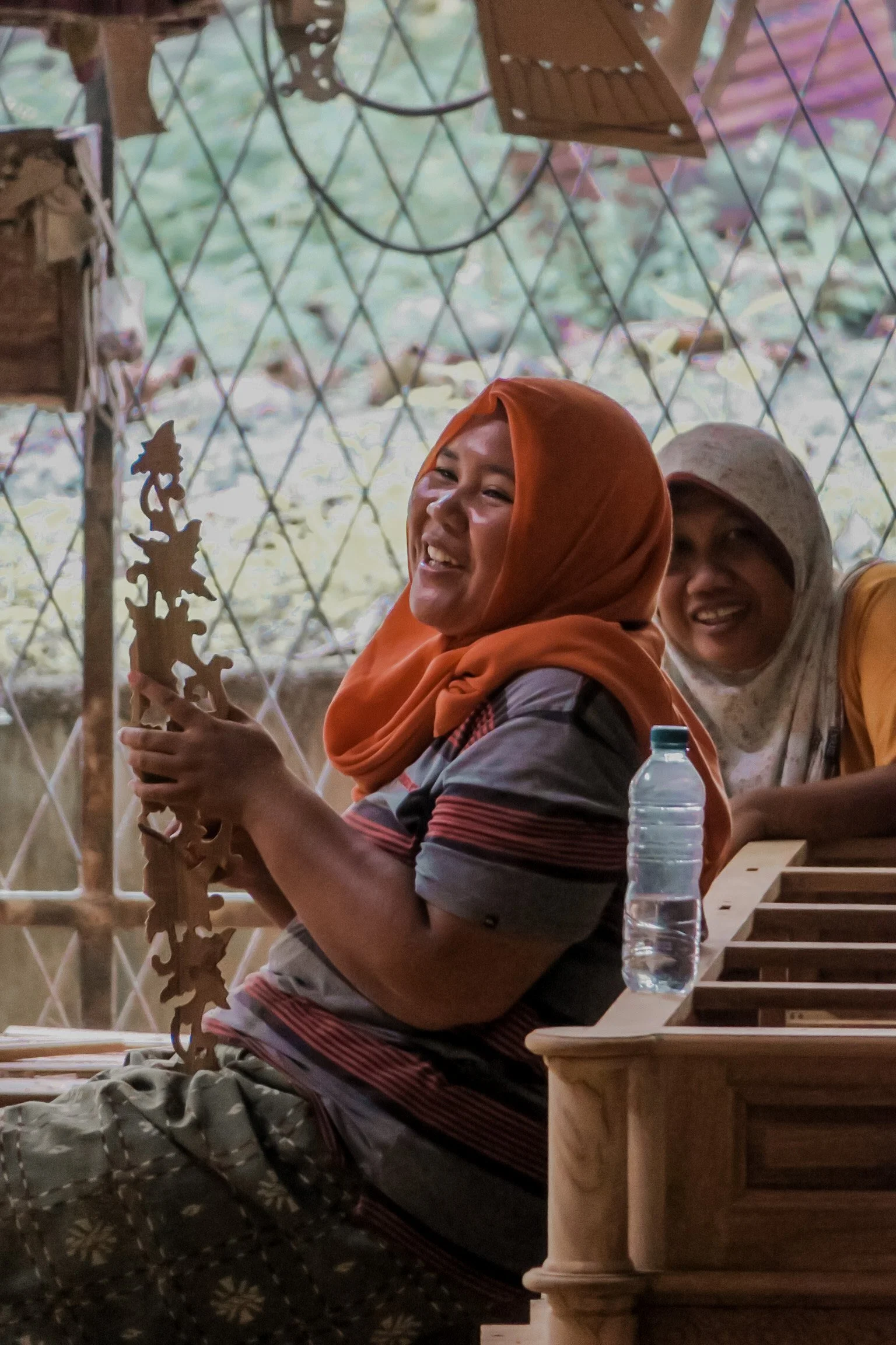 Smiling woman wearing an orange headscarf and striped shirt, sitting at a wooden table with a water bottle, with another smiling woman in the background, Teak Furniture Jepara, wholesale Indonesian teak furniture, custom outdoor teak furniture manufa