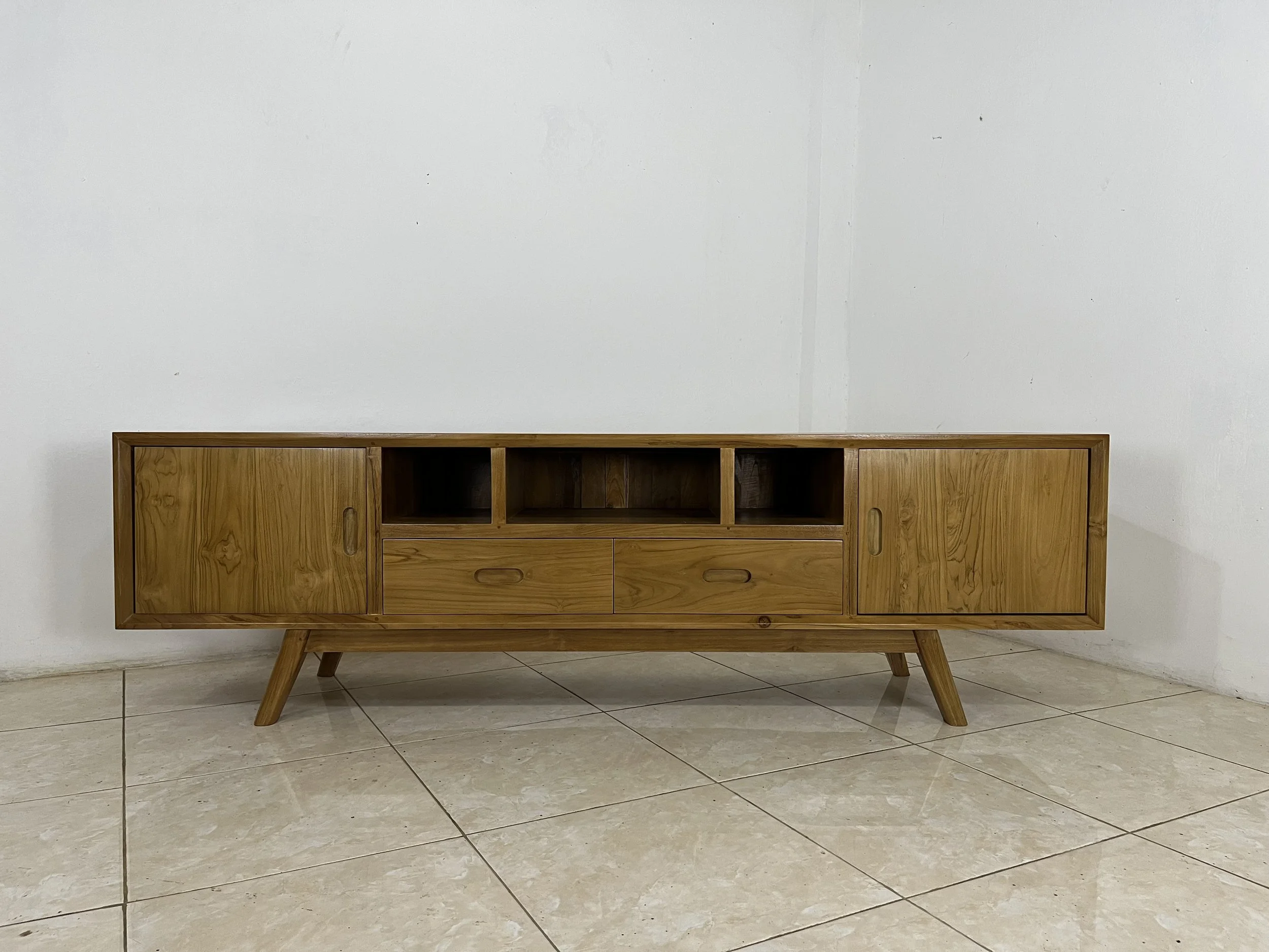 A mid-century modern wooden cabinet with angled legs and multiple storage compartments is placed against a plain white wall on a tiled floor.