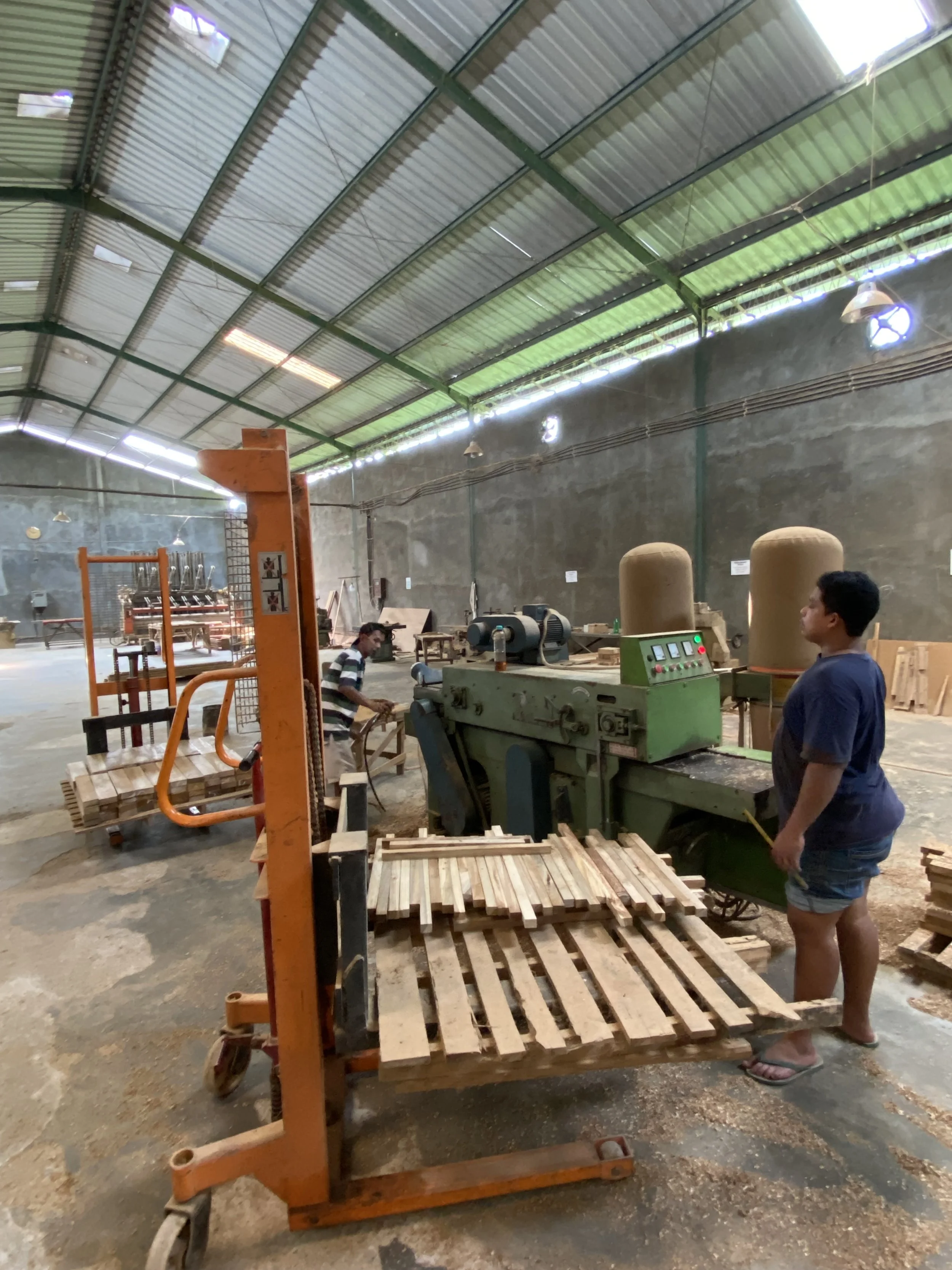 Inside a warehouse or workshop with metal roof and concrete floor, two men working on woodworking equipment surrounded by wooden pallets and materials.