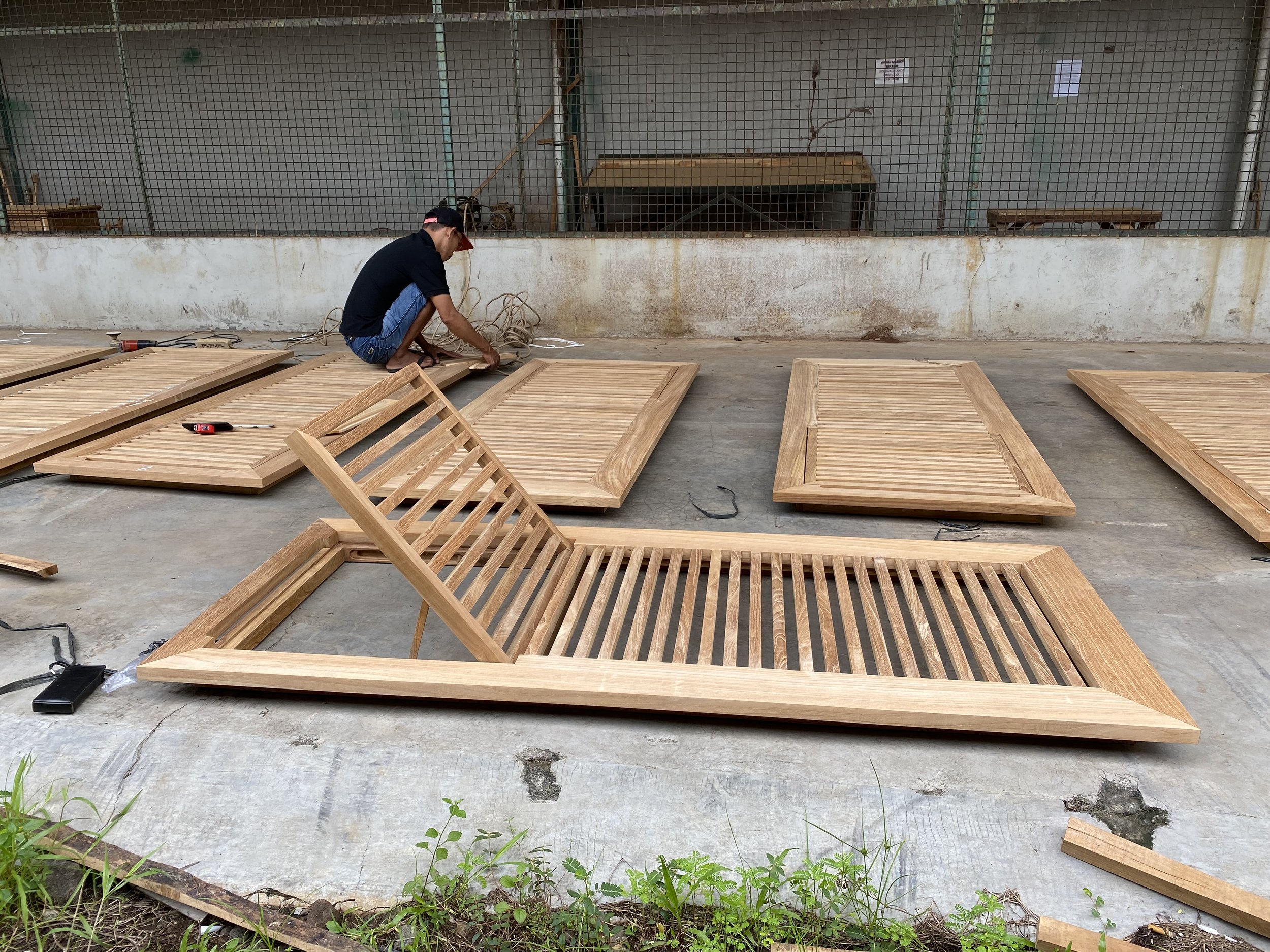 A man working on assembling wooden furniture outdoors on a concrete surface, with several wooden slatted panels laid out, and a wire fence wall behind him.