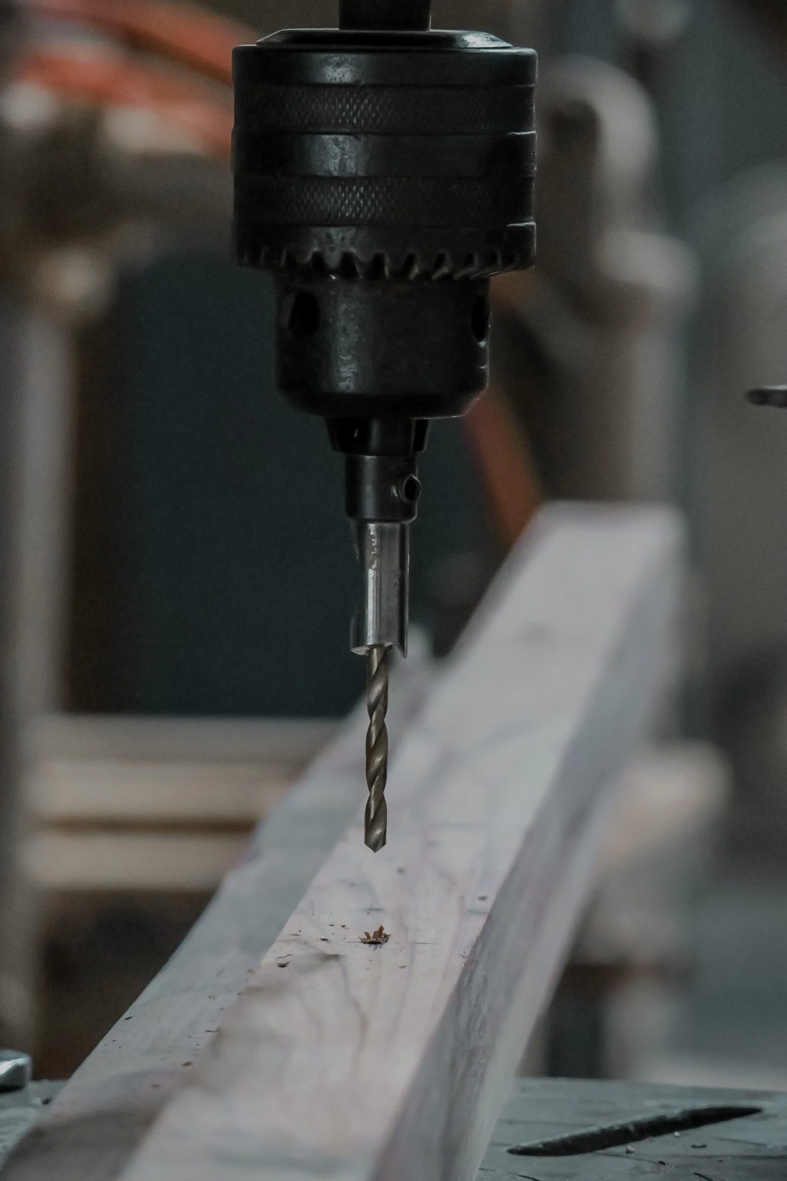 Close-up of a drill bit attached to a drill press, drilling into a wooden surface with sawdust around.