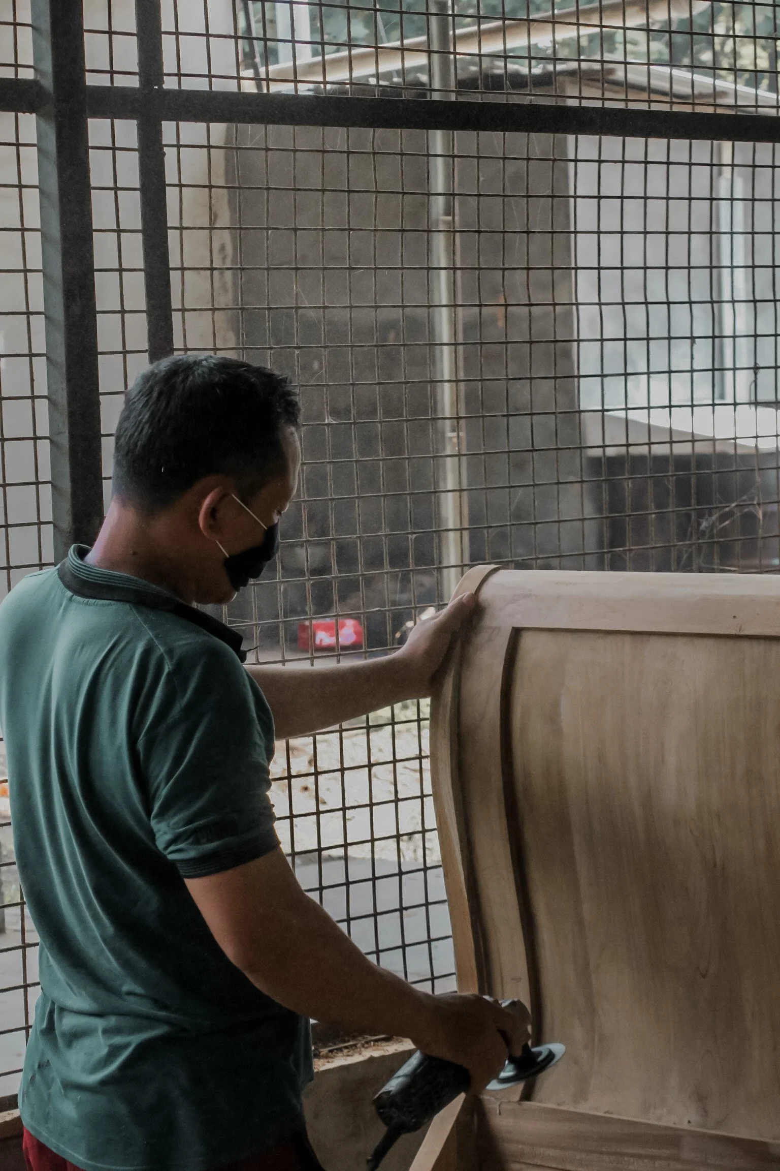 A man wearing a black mask is sanding a wooden piece in an indoor workshop. Teak Furniture Jepara, wholesale Indonesian teak furniture, custom outdoor teak furniture manufacturer, Jepara teak furniture supplier. 
