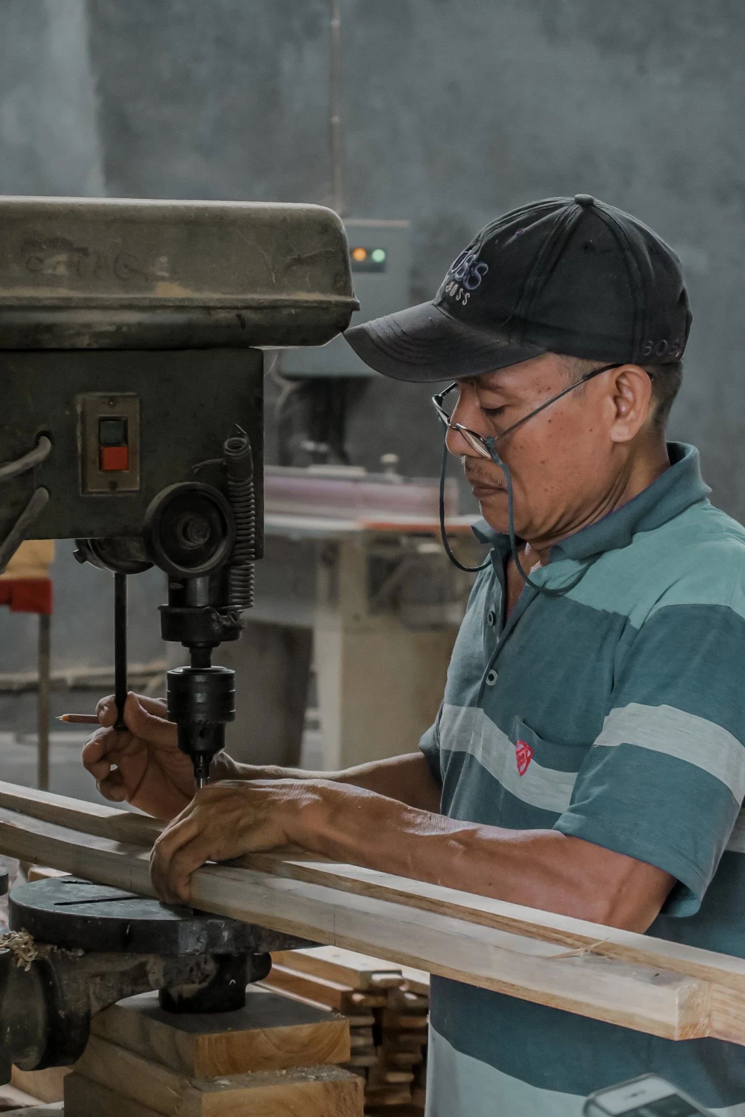 A man in a baseball cap and glasses working with a drill on a wooden piece in a workshop. Teak Furniture Jepara, wholesale Indonesian teak furniture, custom outdoor teak furniture manufacturer, Jepara teak furniture supplier. 