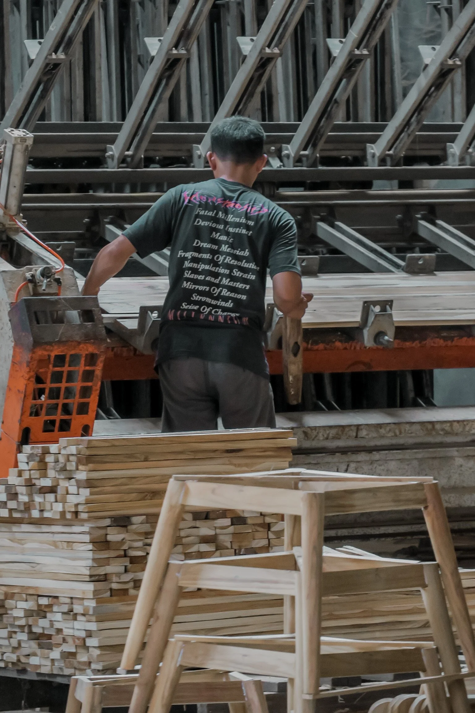 A person working on wooden furniture in a workshop, with stacked wooden pieces and chairs nearby. Teak Furniture Jepara, wholesale Indonesian teak furniture, custom outdoor teak furniture manufacturer, Jepara teak furniture supplier. 