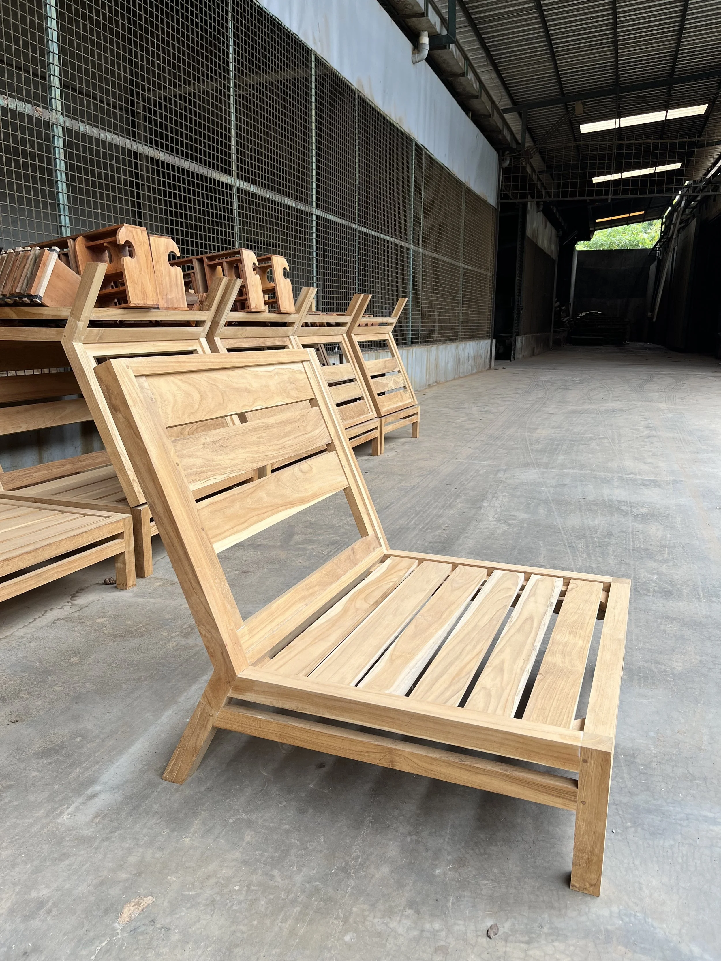 Stacked wooden chairs in an industrial setting with a metal grid wall and a concrete floor.