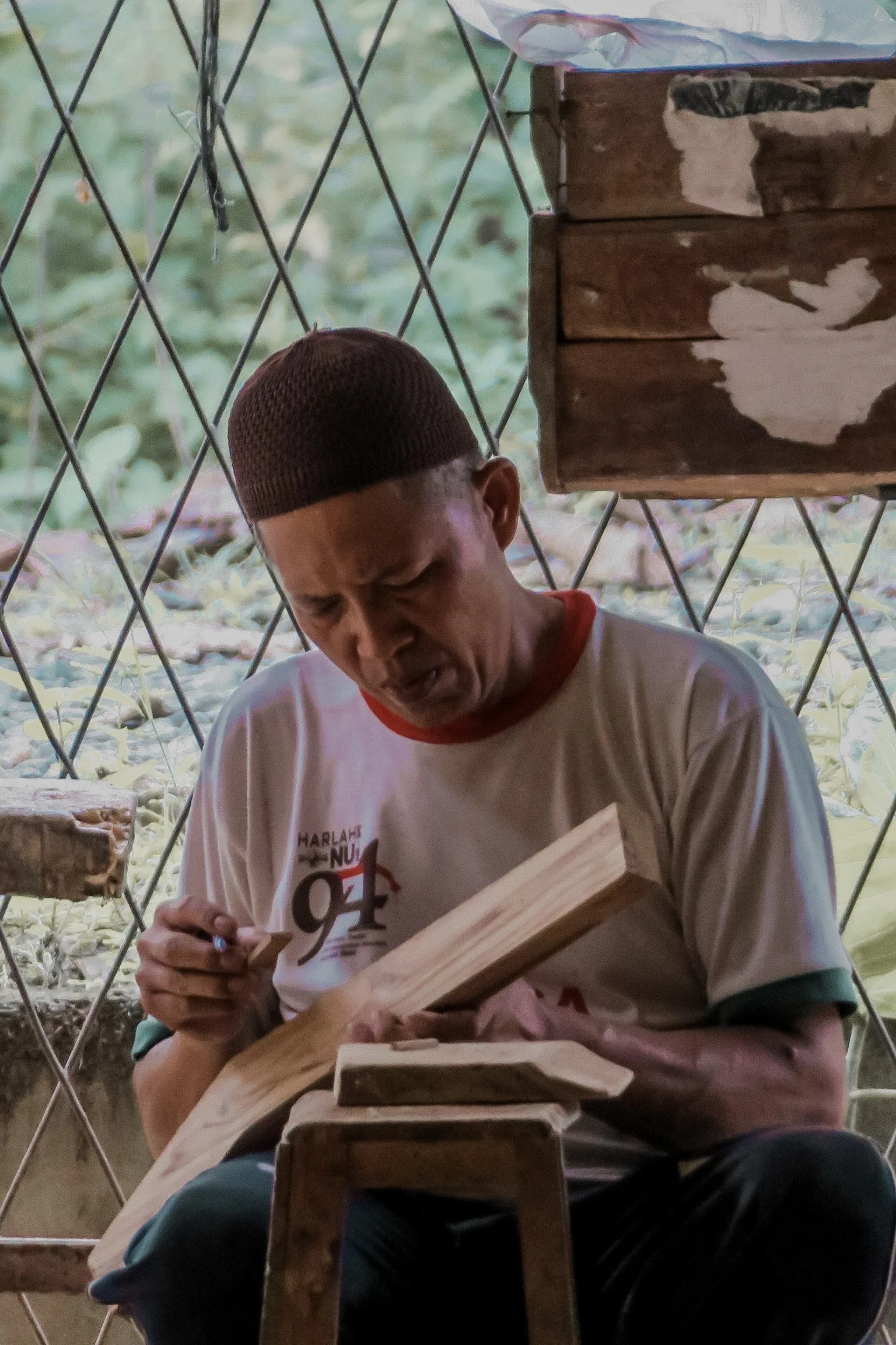 A man wearing a brown knit cap and a white T-shirt with red and black text is sitting at a table, signing a wooden object with a pen. Teak Furniture Jepara, wholesale Indonesian teak furniture, custom outdoor teak furniture manufacturer, Jepara teak 