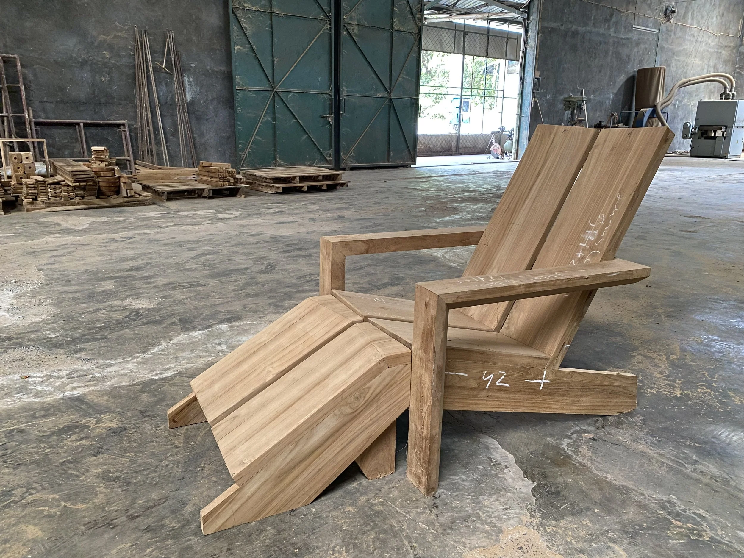 Industrial workshop with wooden furniture pieces, including a reclining chair and various wood planks, inside a large workshop with open sliding doors, concrete floor, and metal walls.