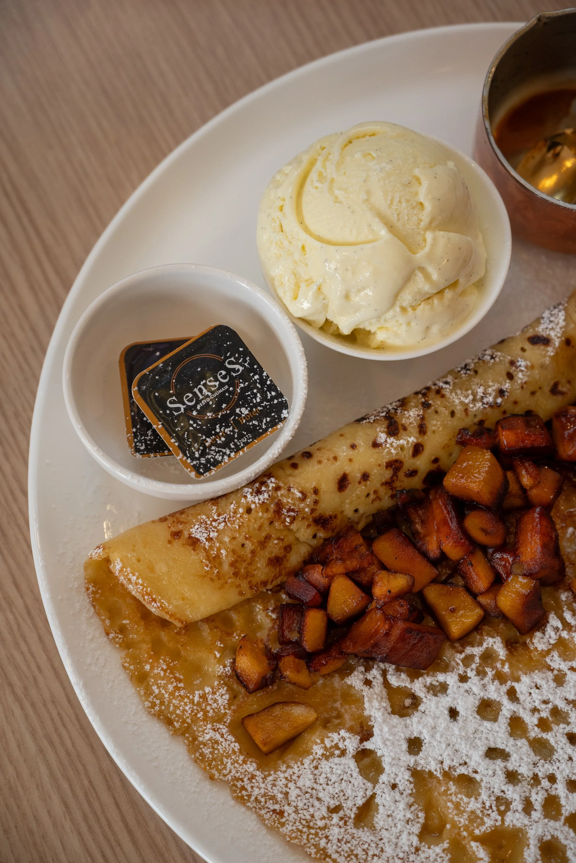 A white plate with a thin crepe topped with caramelized fruit pieces, a scoop of vanilla ice cream in a small dish, and a chocolate-covered jelly dessert with powdered sugar, served with a small cup of chocolate sauce.