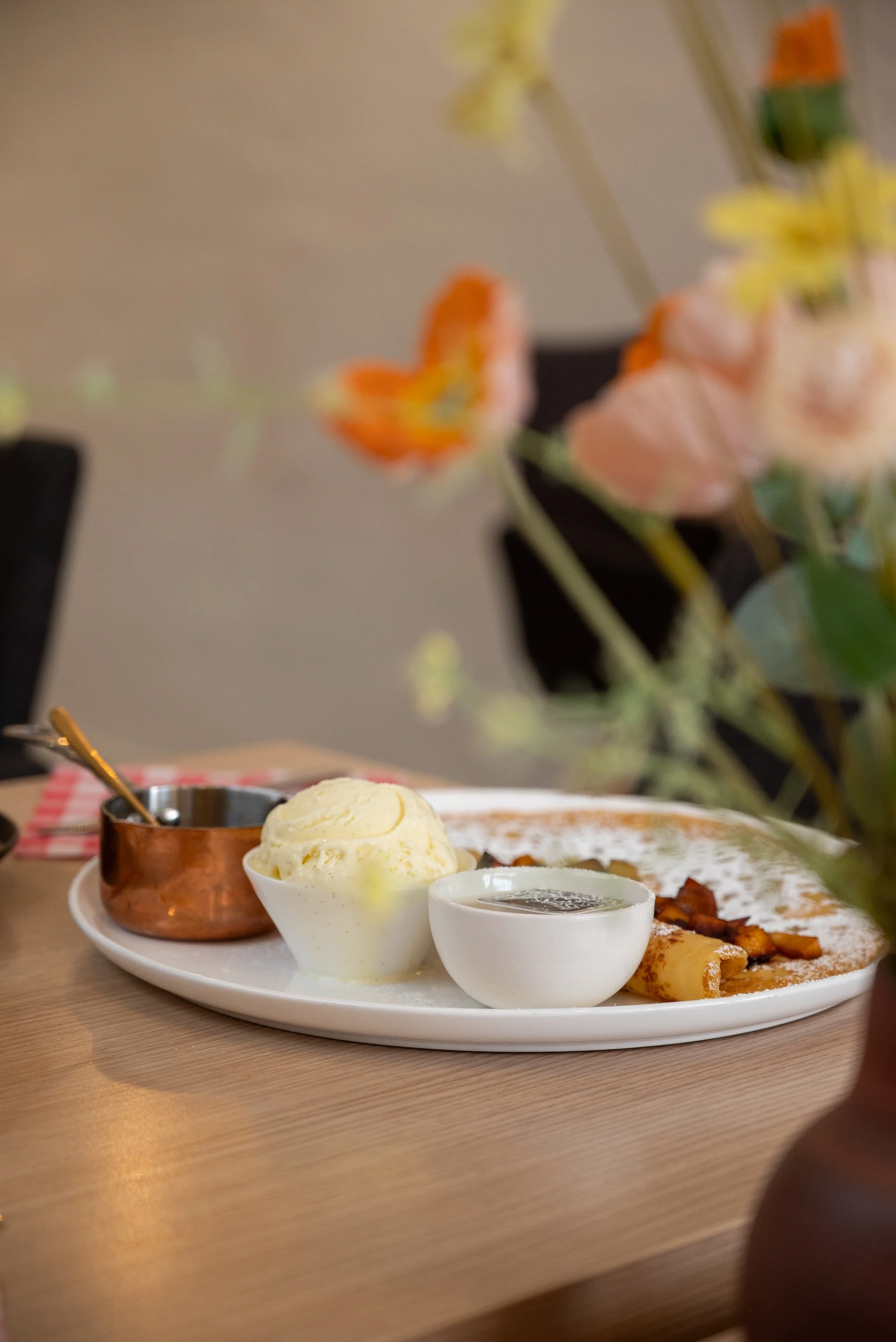 A breakfast plate with a scoop of vanilla ice cream, a small bowl of syrup, a small copper pot, and a stack of pancakes with bacon on a white plate, set on a wooden table.