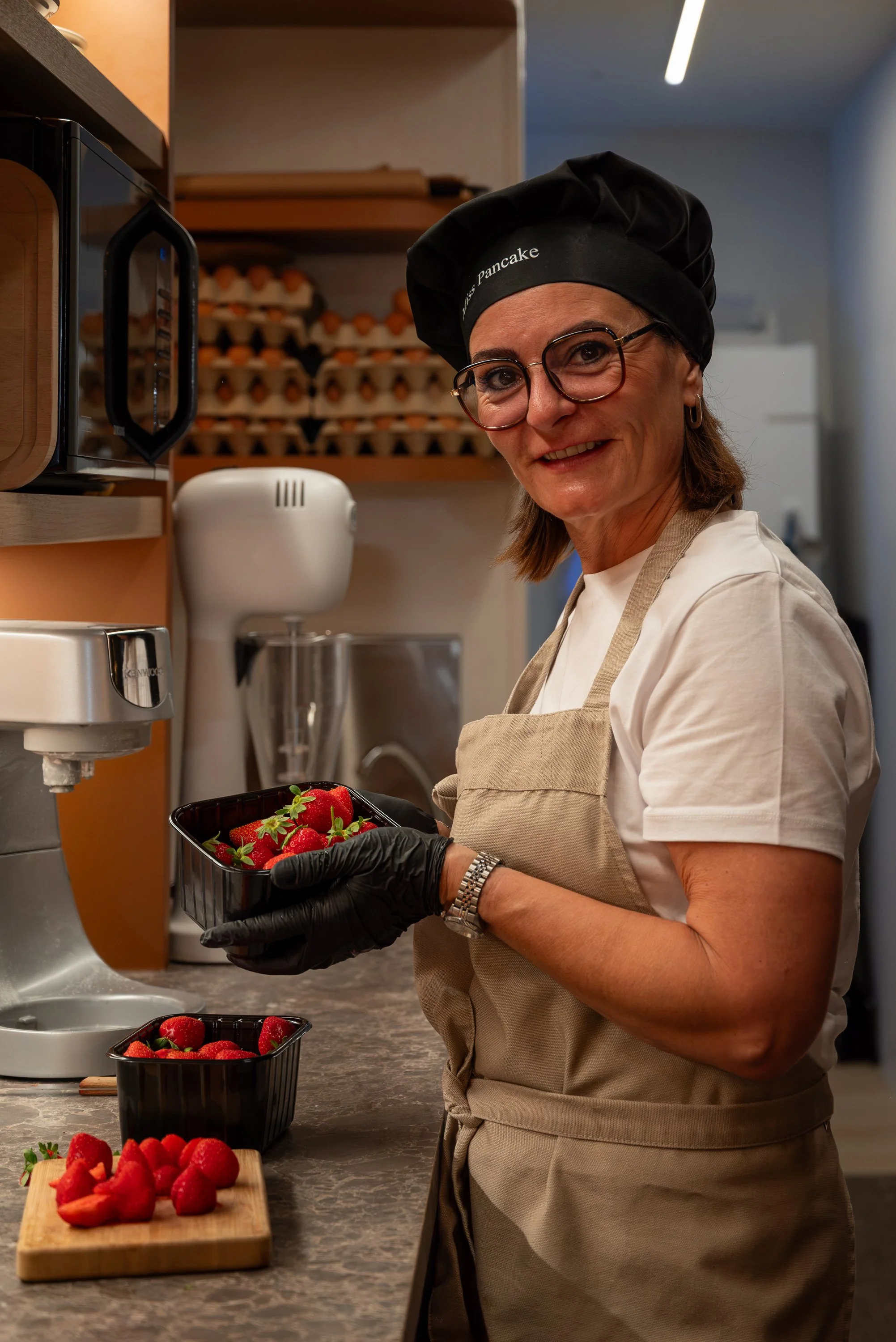 A woman wearing glasses, a black chef hat, a white shirt, and a beige apron holds a black container of strawberries in a kitchen.