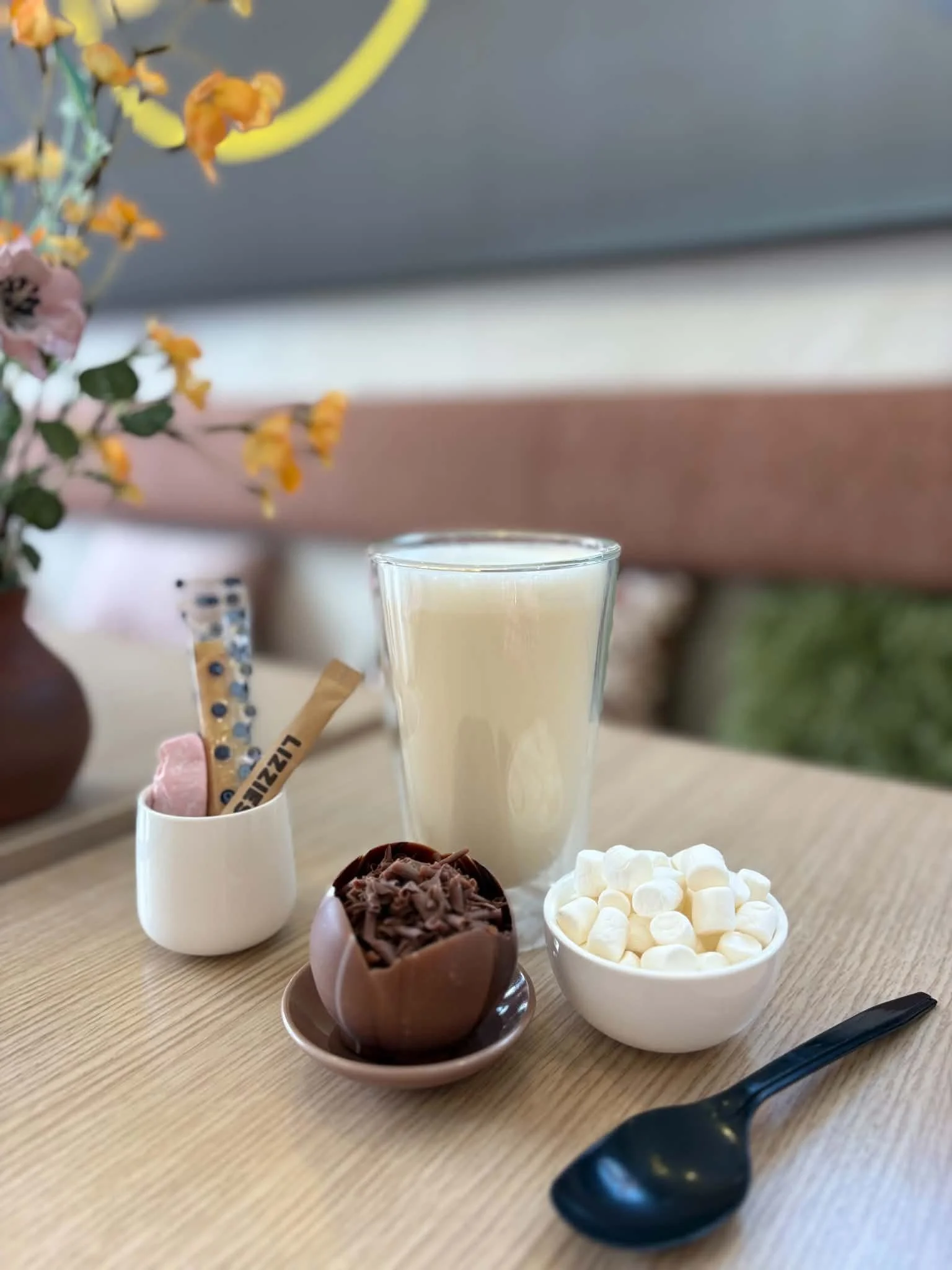 A glass of vanilla milkshake on a wooden table with bowls of mini marshmallows, chocolate chips, and ice cream, and a spoon nearby.
