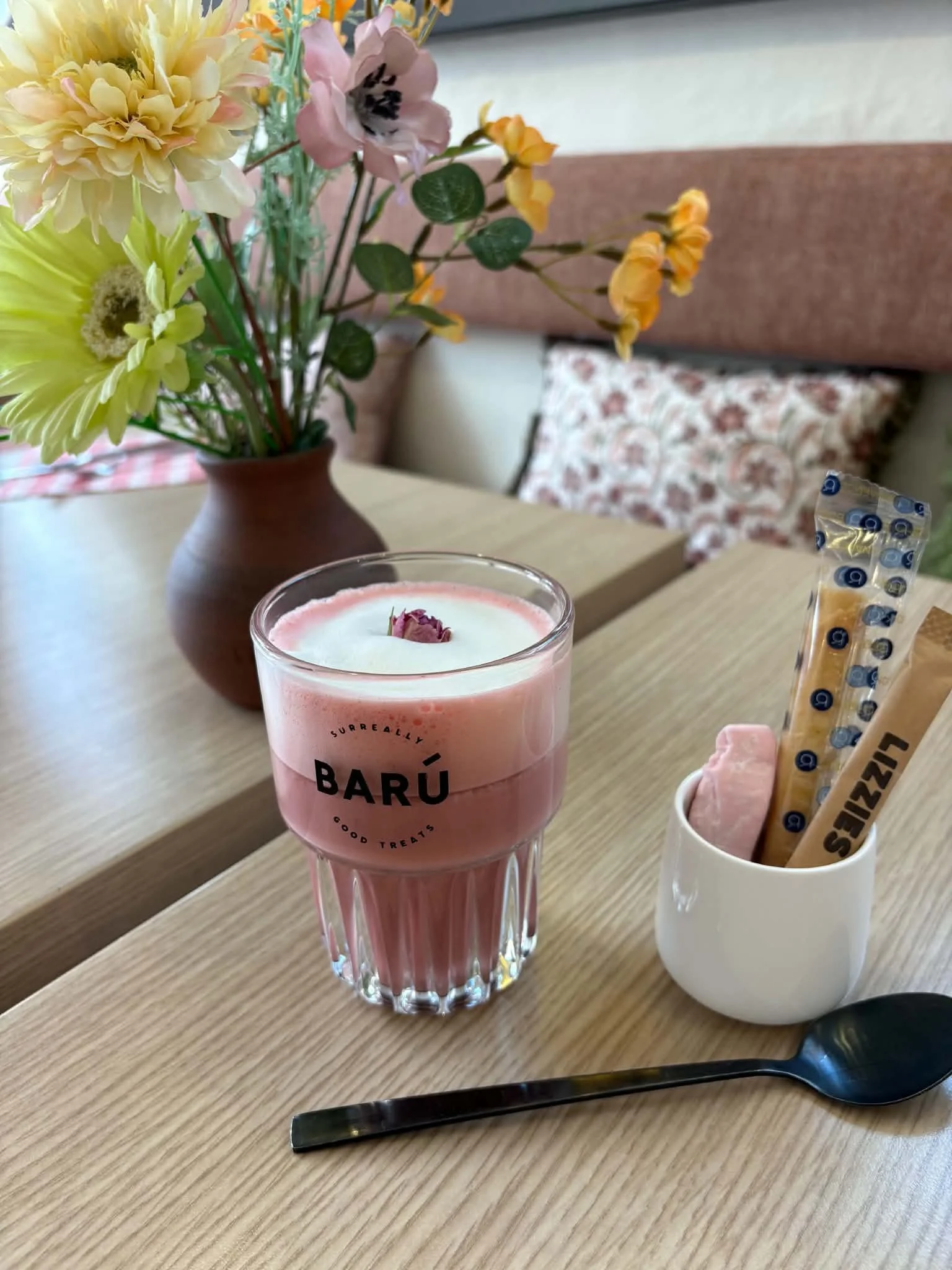 A pink candle with a dried rose on top is on a wooden table. Next to it, there's a white cup holding pink & white candies, and a decorative knife. In the background, a vase with yellow, pink, and purple flowers is on a wooden table, with a floral cus