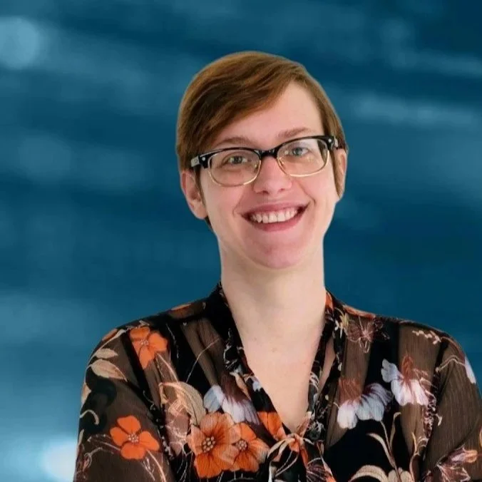 A woman with short brown hair, glasses, and a floral blouse, smiling at the camera against a blue background.