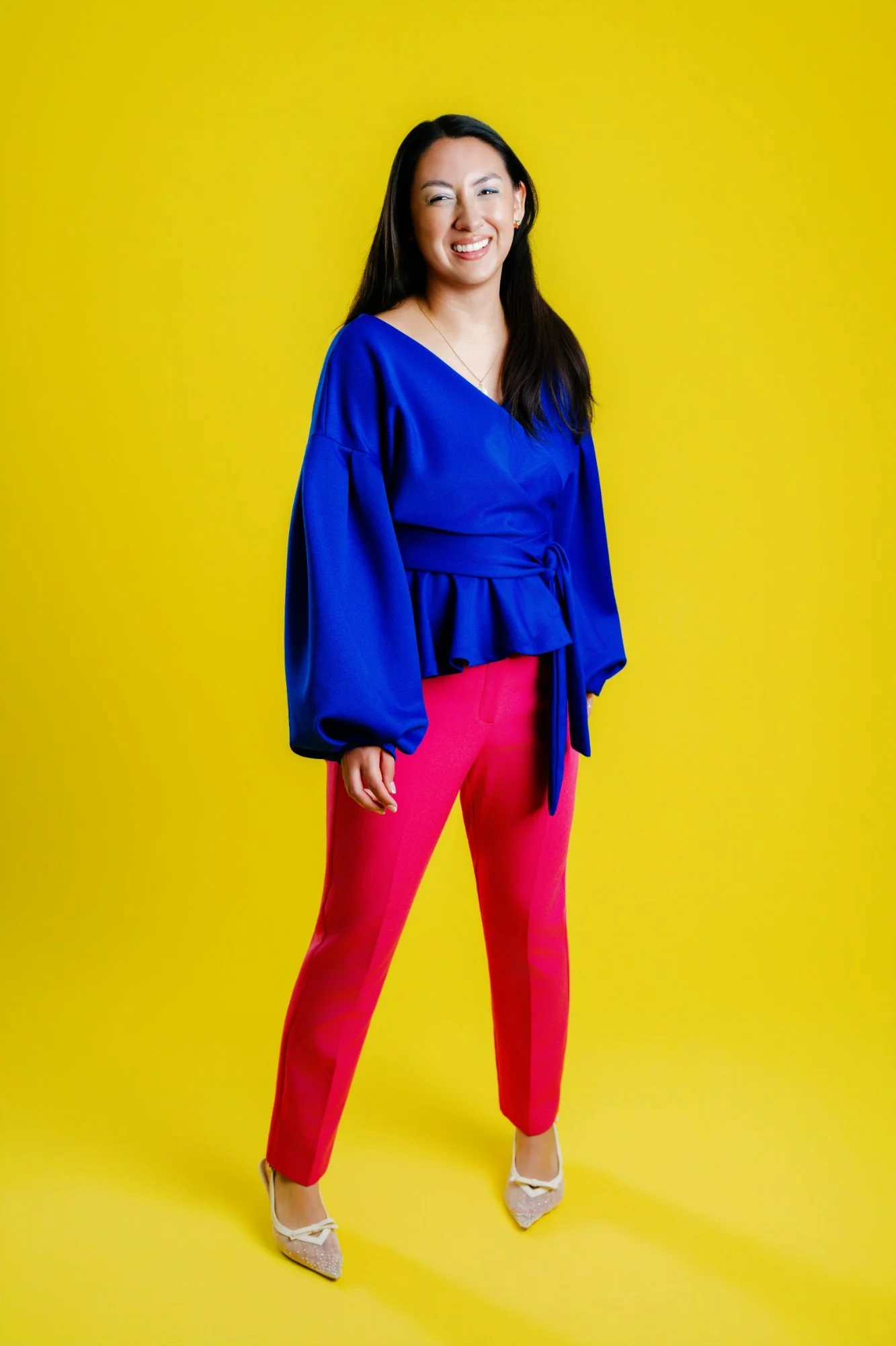 A woman with long dark hair wearing a blue top, pink pants, white shoes, and jewelry standing against a yellow background.