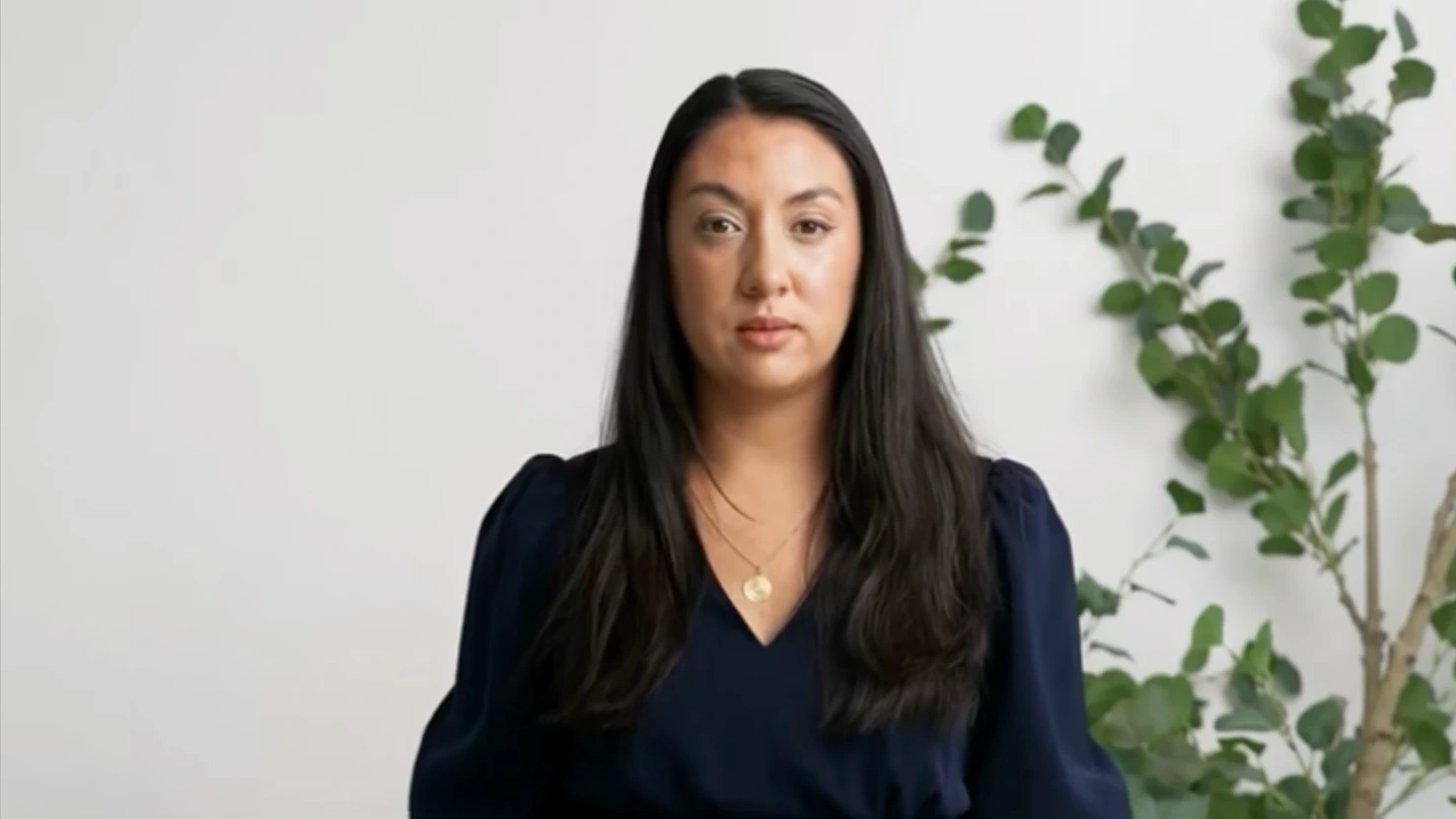 A woman with long black hair, wearing a dark blue top and a gold necklace, is sitting in front of a white wall and a green plant.