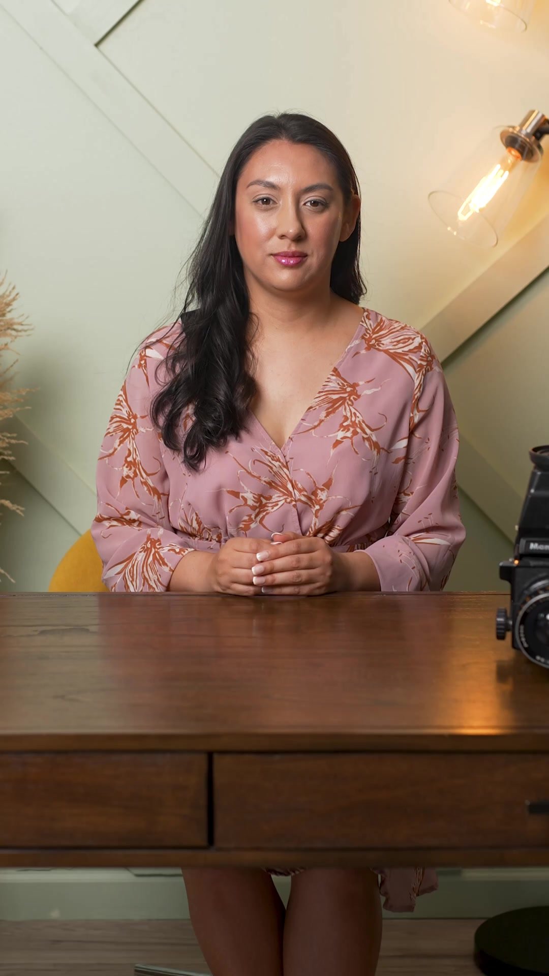 A woman with long dark hair, wearing a pink floral satin robe, sits at a wooden table with her hands clasped, in a room with a modern lightbulb on the wall and a camera on the table.