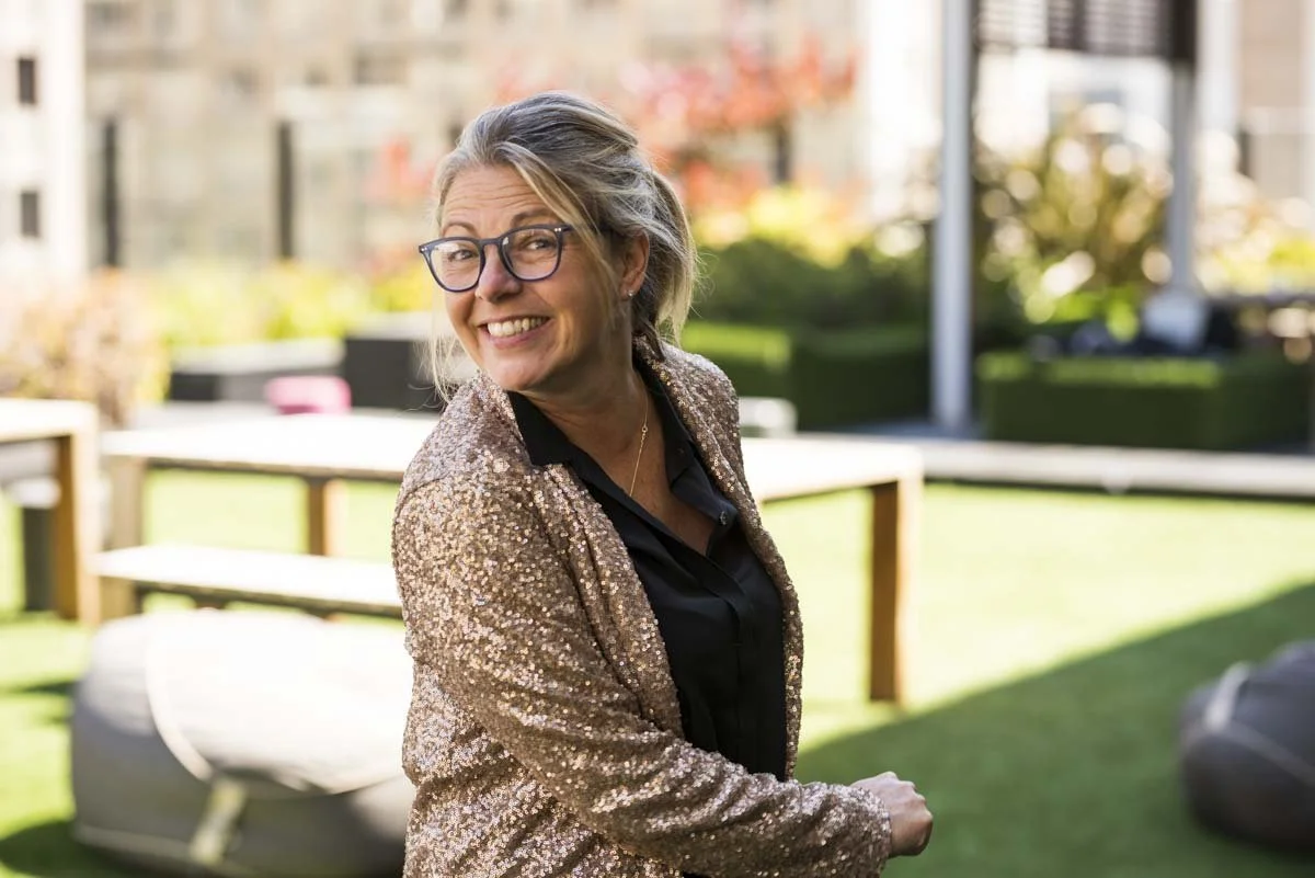 A woman with gray hair and glasses smiling outdoors in a sunny, landscaped area.