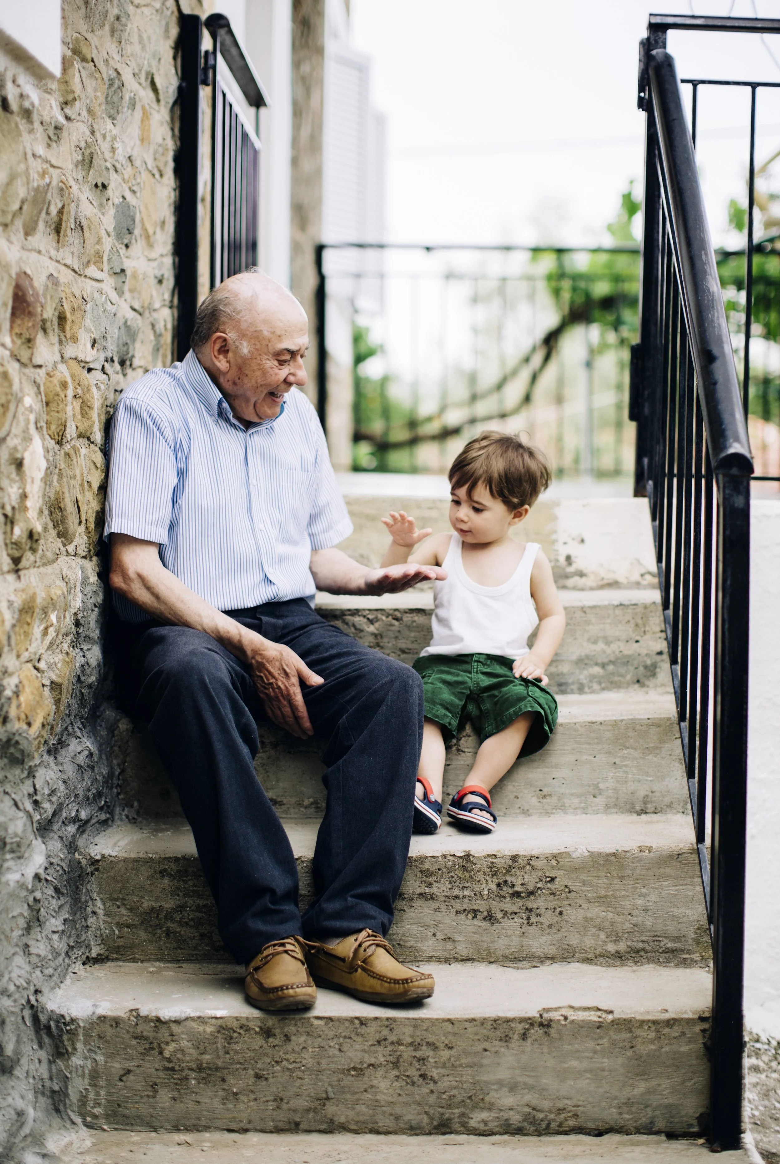 An elderly man and a young boy sitting on outdoor concrete stairs, talking and smiling at each other. The man is wearing a blue striped shirt and dark pants, the boy is in a white tank top, green shorts, and sandals. There is a stone wall on the left and a black railing on the right.