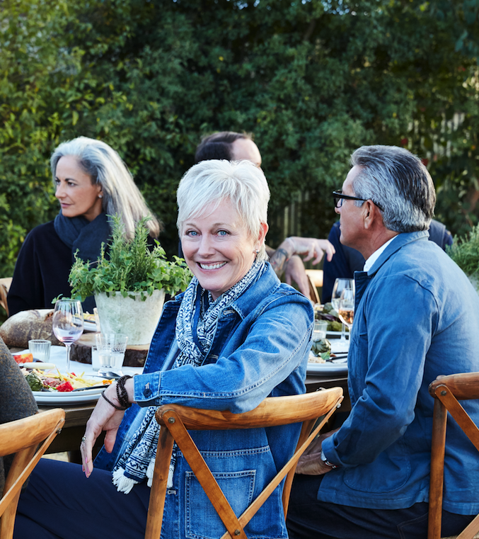 People enjoying an outdoor dinner party with food and drinks, seated around a table with greenery in the background.