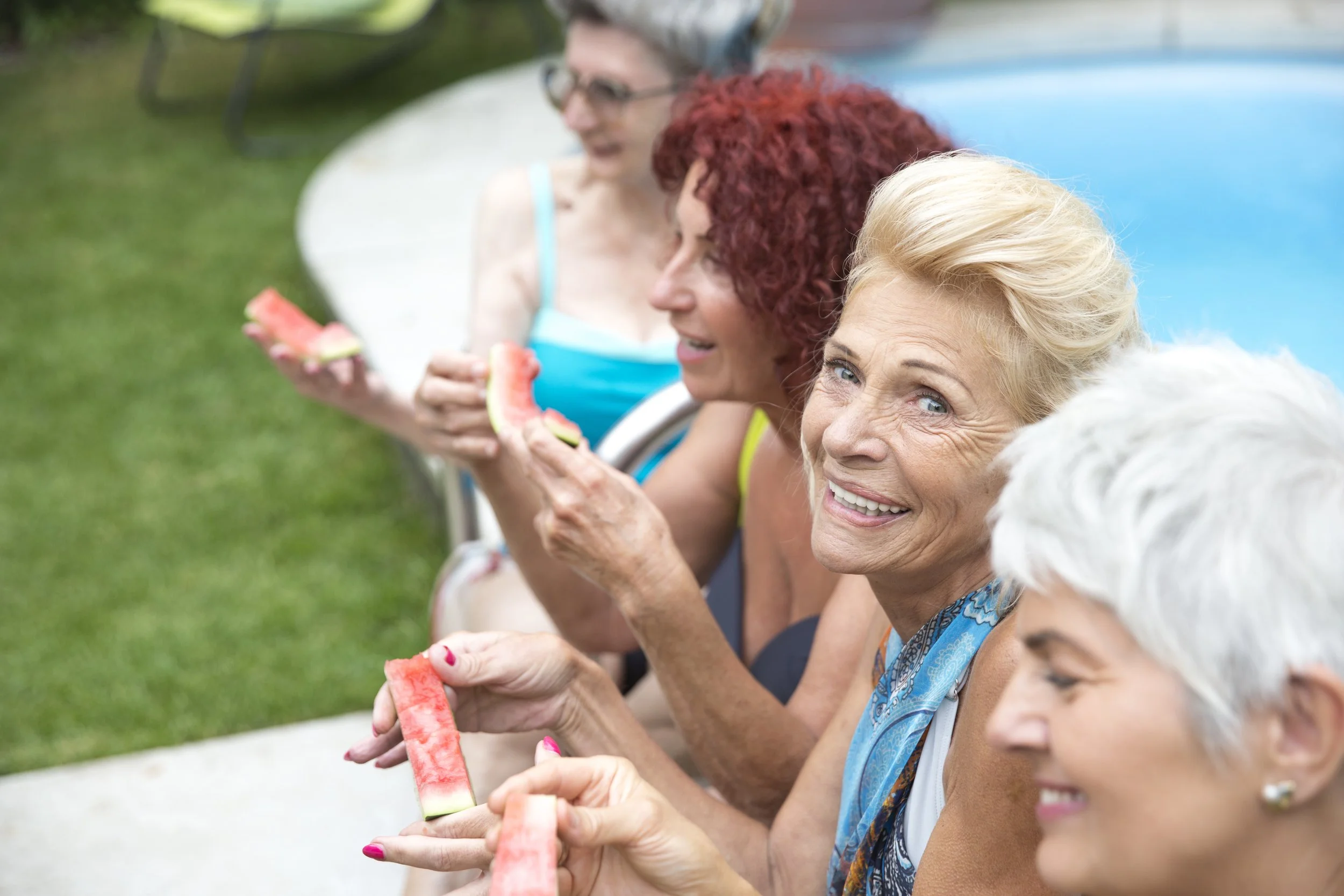 Group of women sitting outdoors, eating watermelon slices, smiling and enjoying a sunny day.