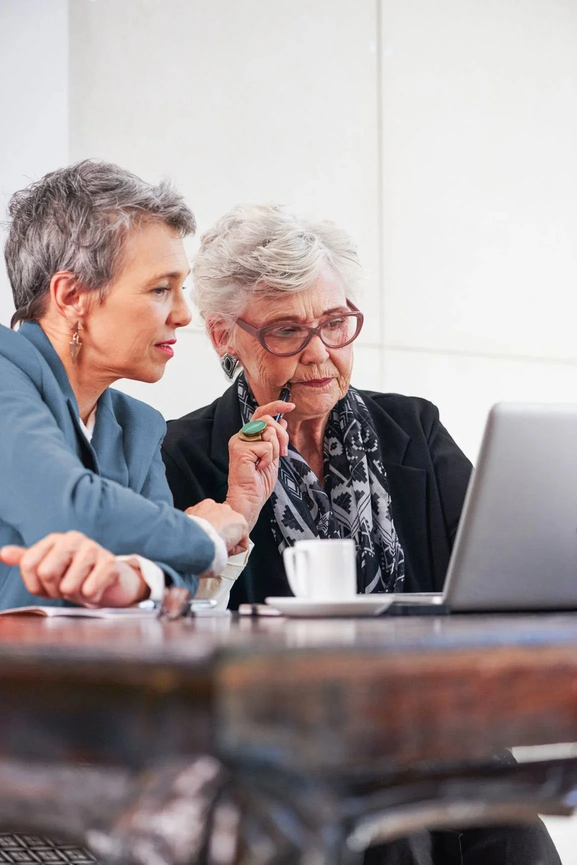Two women, one with gray hair and glasses and one with short gray hair, sit at a table looking at a laptop. The woman with glasses holds a pen near her mouth. There are a white cup and saucer on the table.