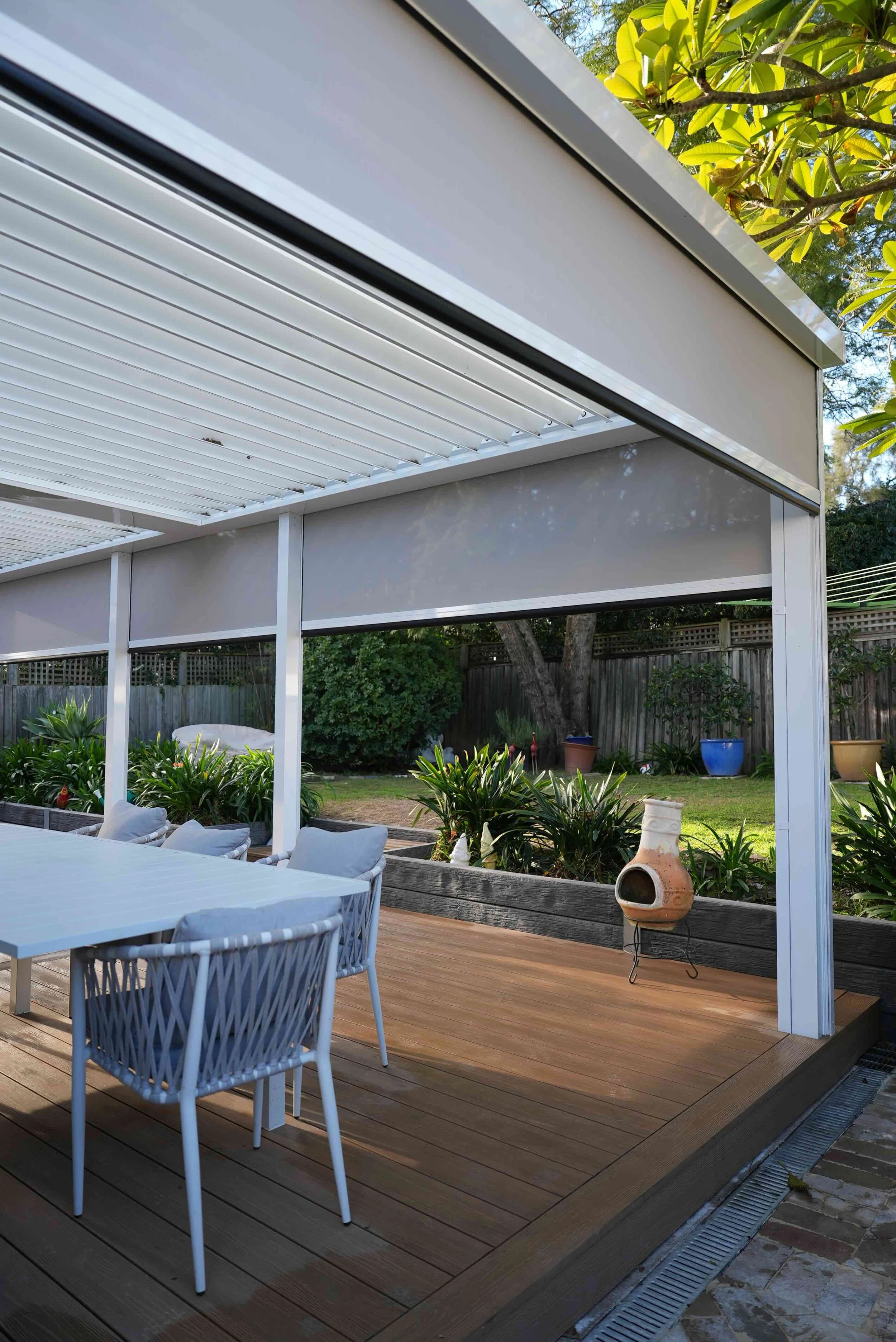 An outdoor patio with a wooden deck, white dining table, and chairs, surrounded by potted plants and greenery, with a modern white metal pergola overhead.