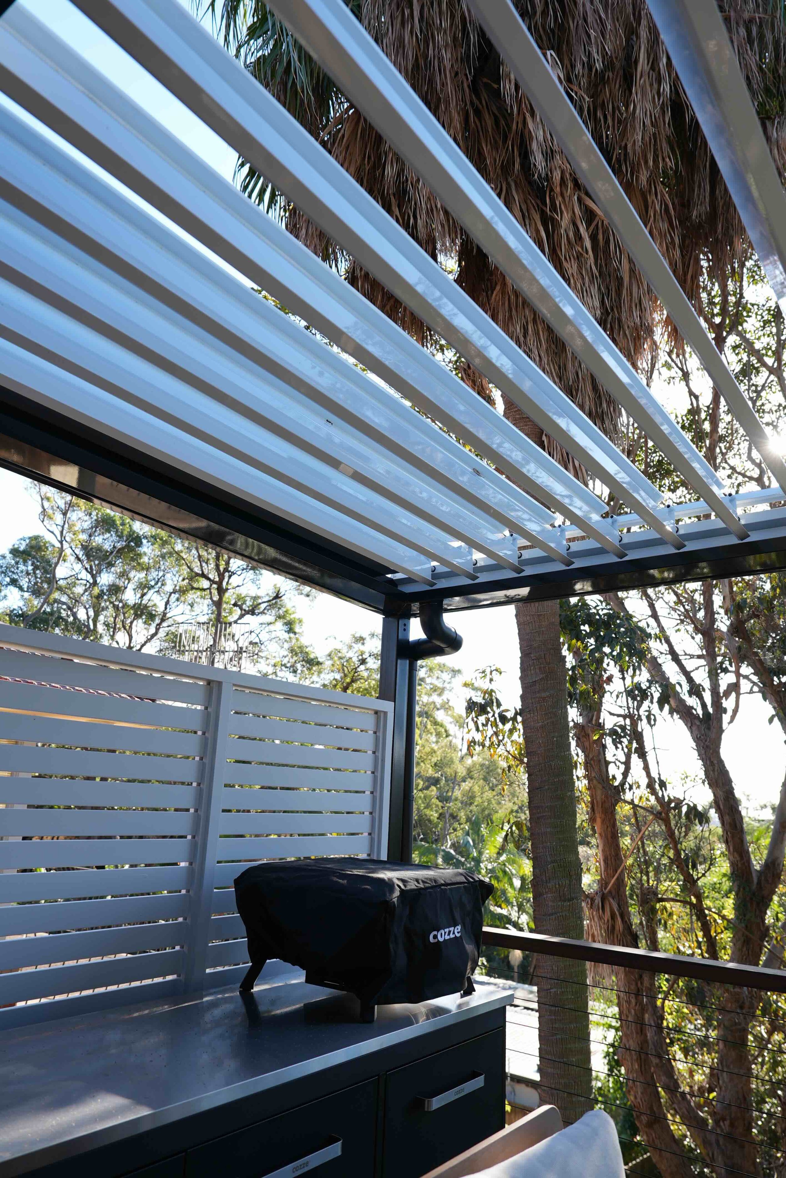 Outdoor balcony or deck area with white slatted railing, trees visible in the background, and a sunlit sky. On the table, there is a black object covered with a protective cover.