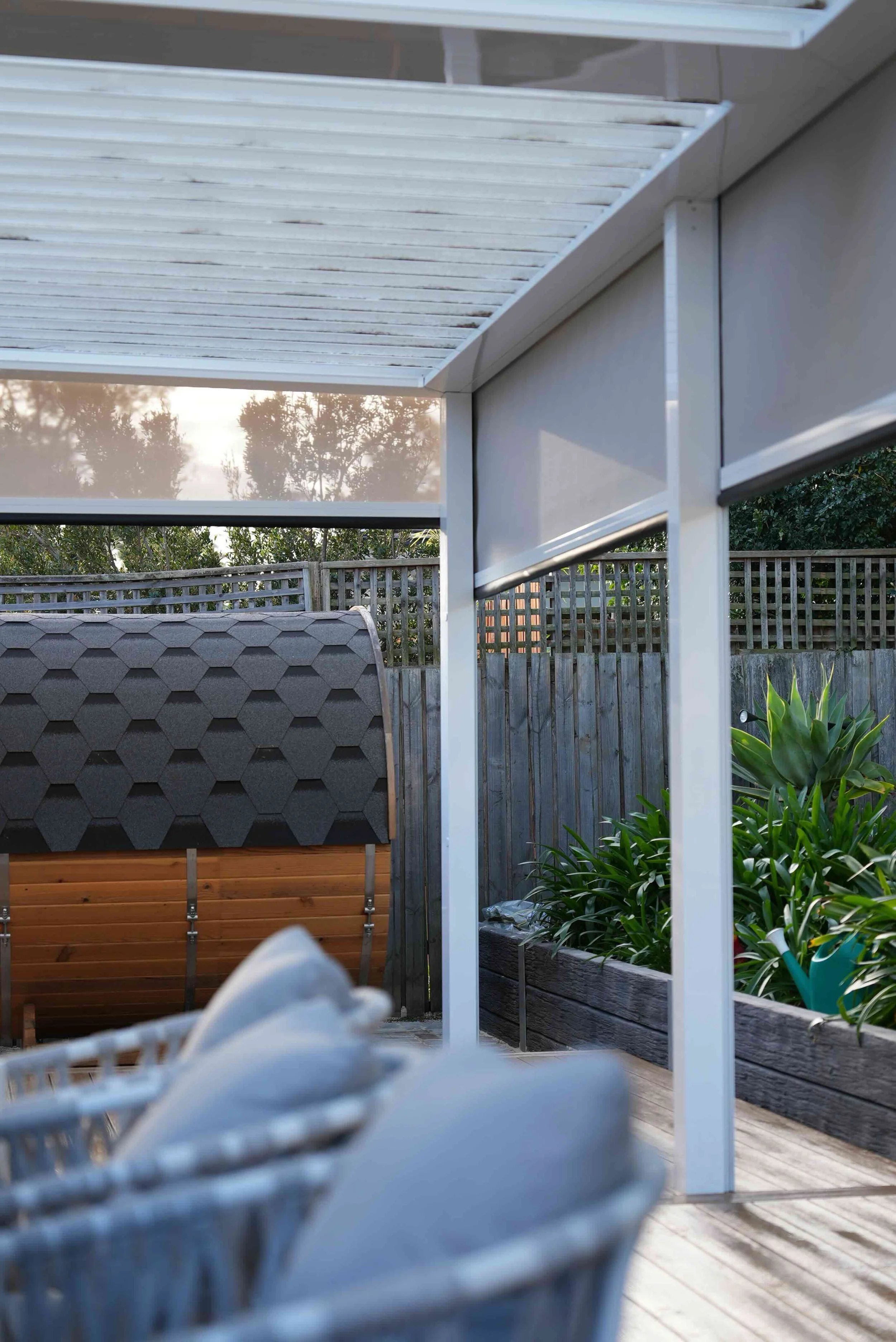 View of a backyard patio area with a white pergola, a wooden fence, a hedge, and plants in a garden bed.