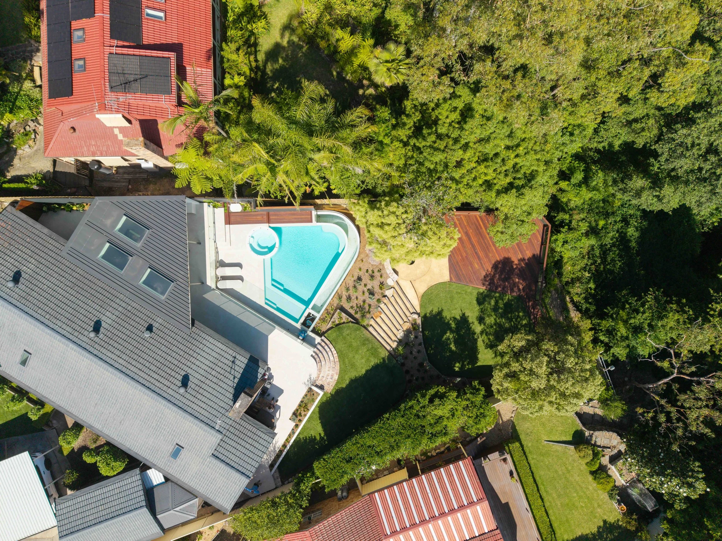 An aerial view of a residential backyard area featuring a swimming pool with a curved shape, surrounded by a patio, green grass, and various trees and plants. Part of a house with a gray roof and skylights is visible, as well as neighboring houses wi