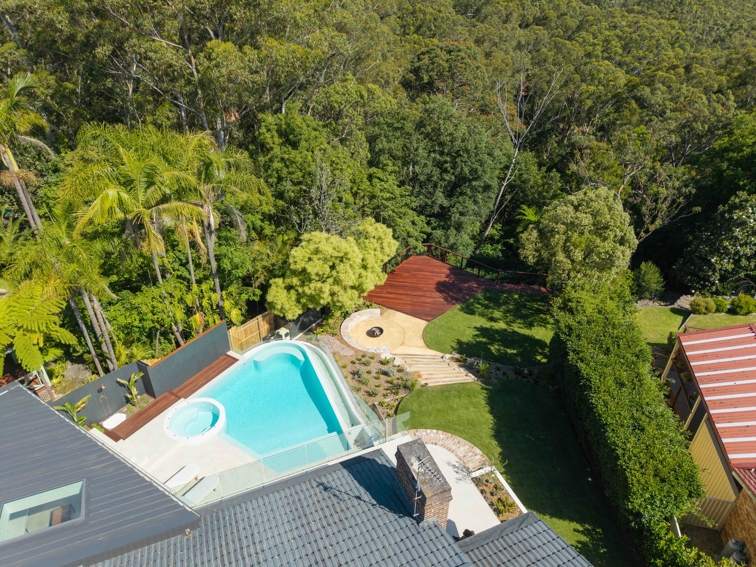 A bird's-eye view of a backyard with a swimming pool, a hot tub, a wooden deck, and a grassy lawn, surrounded by dense trees and greenery.