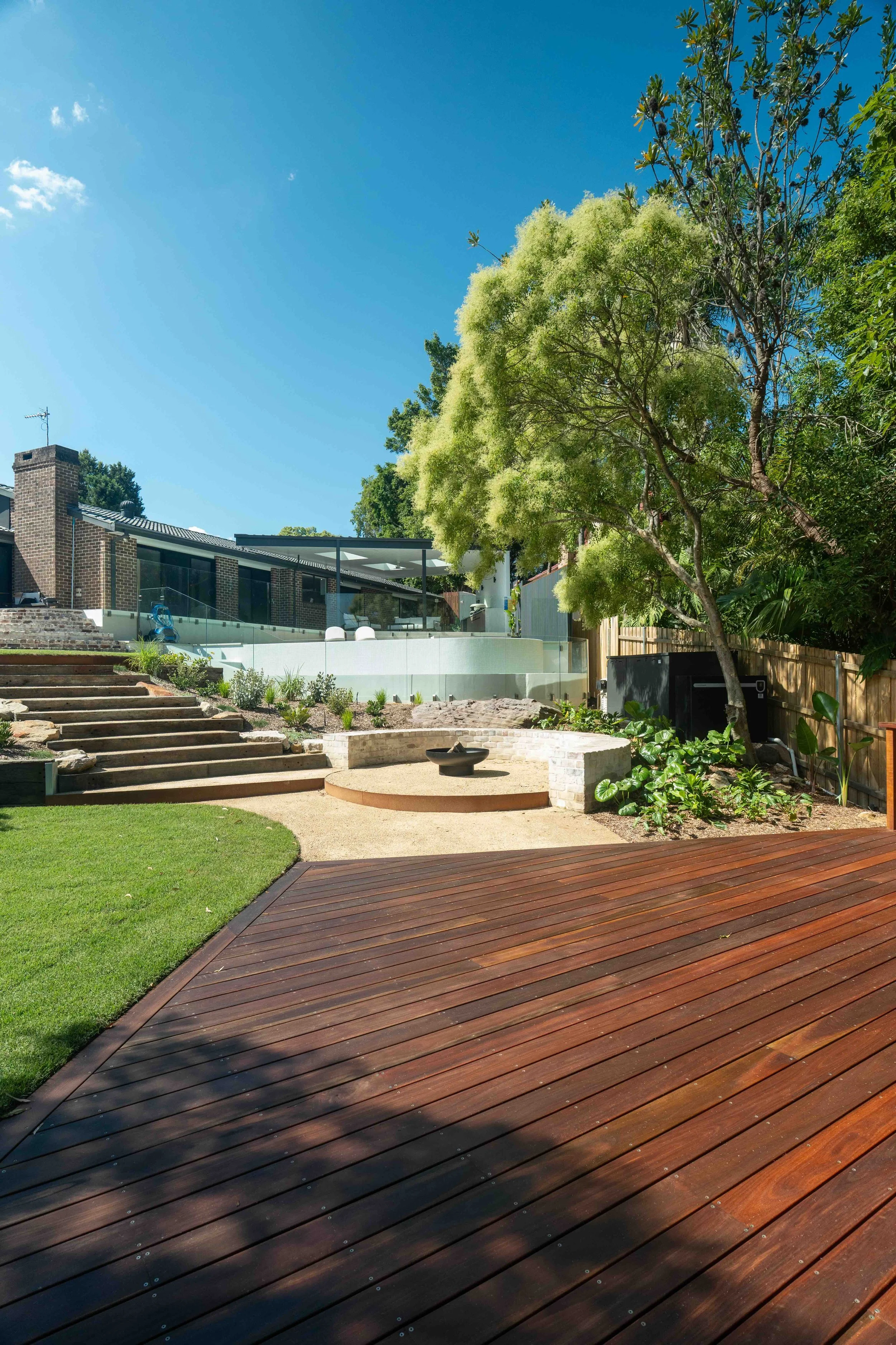 View of a backyard with wooden deck, green lawn, stone steps, a tree with light green foliage, and a modern house with glass walls and a patio. The sky is clear with a few clouds.