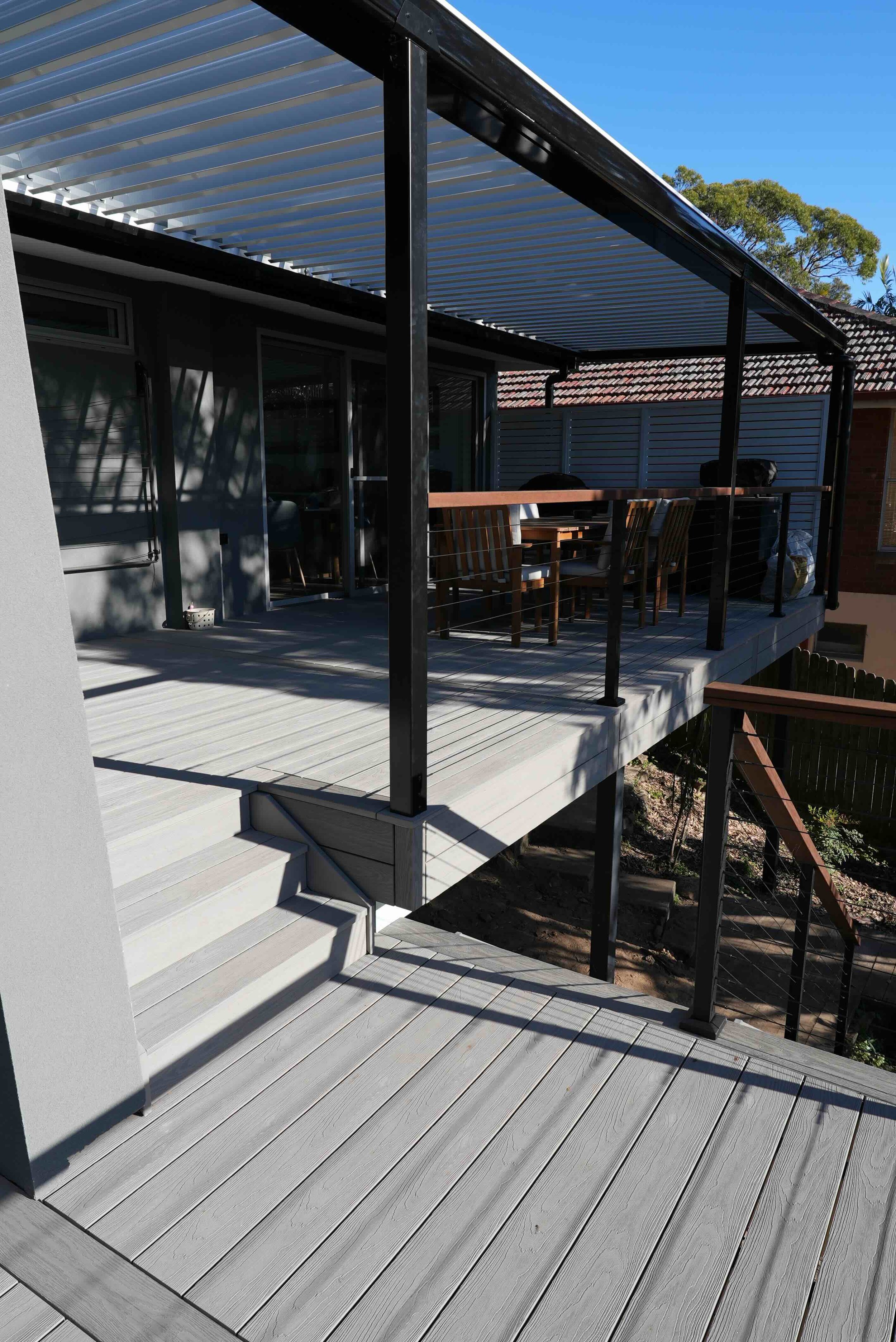 A modern outdoor patio with light gray composite decking, black metal railing, and a roof with horizontal slats, attached to a house with sliding glass doors and a brick exterior.
