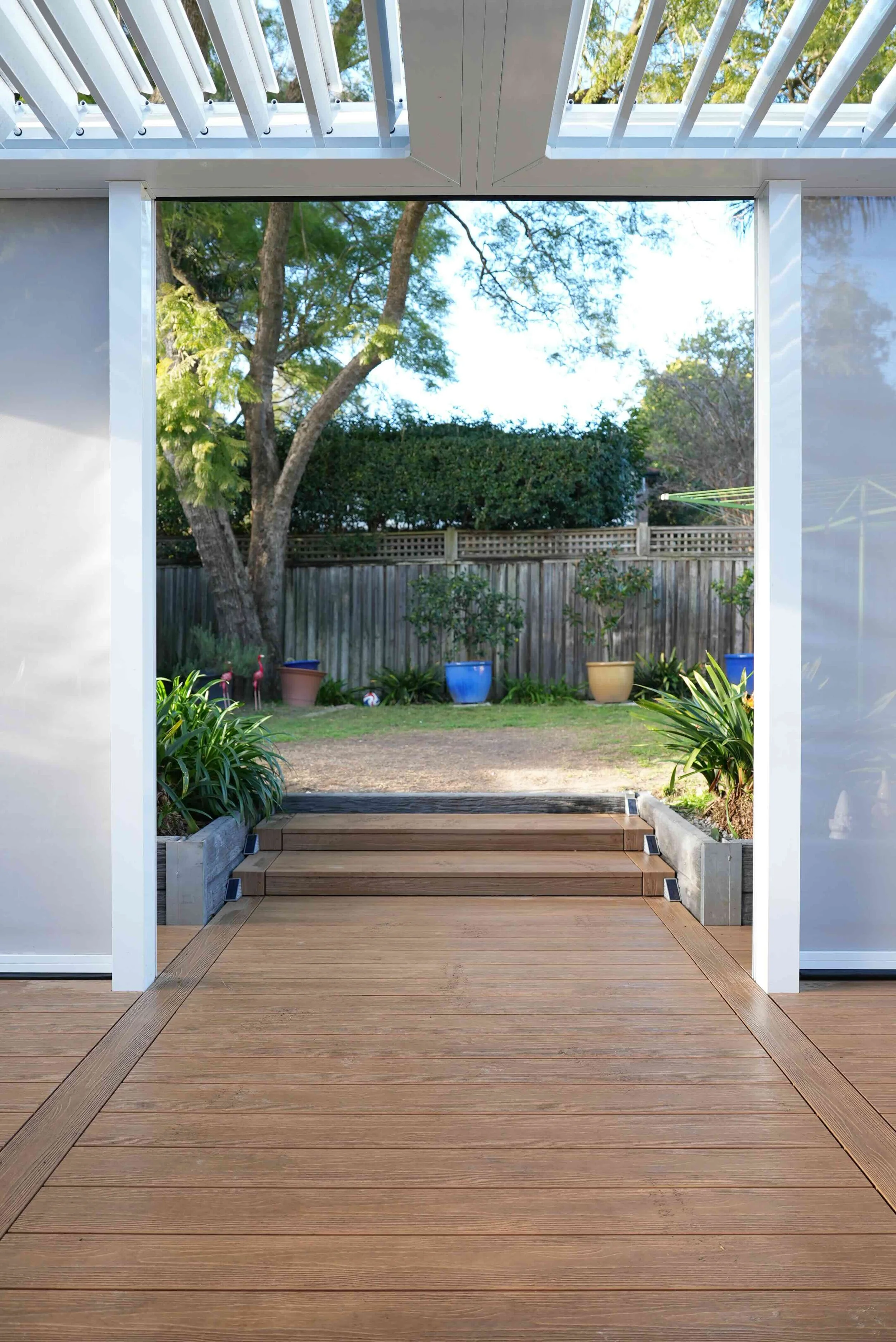View from a covered patio with wooden steps leading to a backyard with potted plants, trees, and a wooden fence.