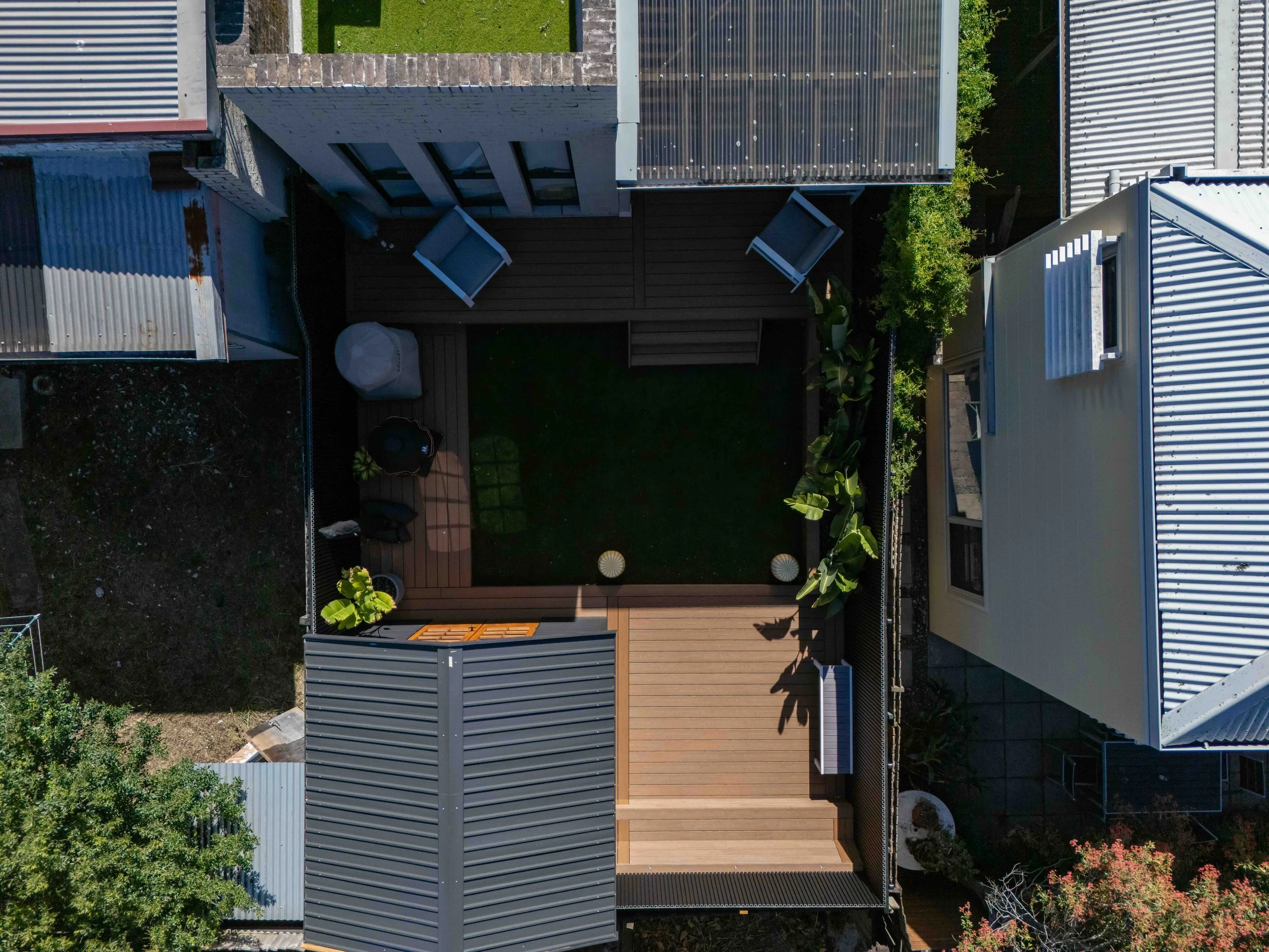 Bird's eye view of rooftops with various metal roofing panels, some with corrugated patterns, and a small backyard area with patio furniture and landscaping.