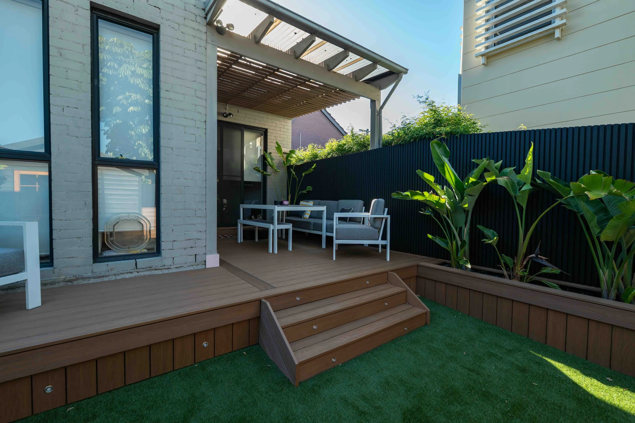 Backyard patio with wooden steps, patio furniture including a table and cushioned chairs, green plants, and a black privacy fence under a partly cloudy sky.