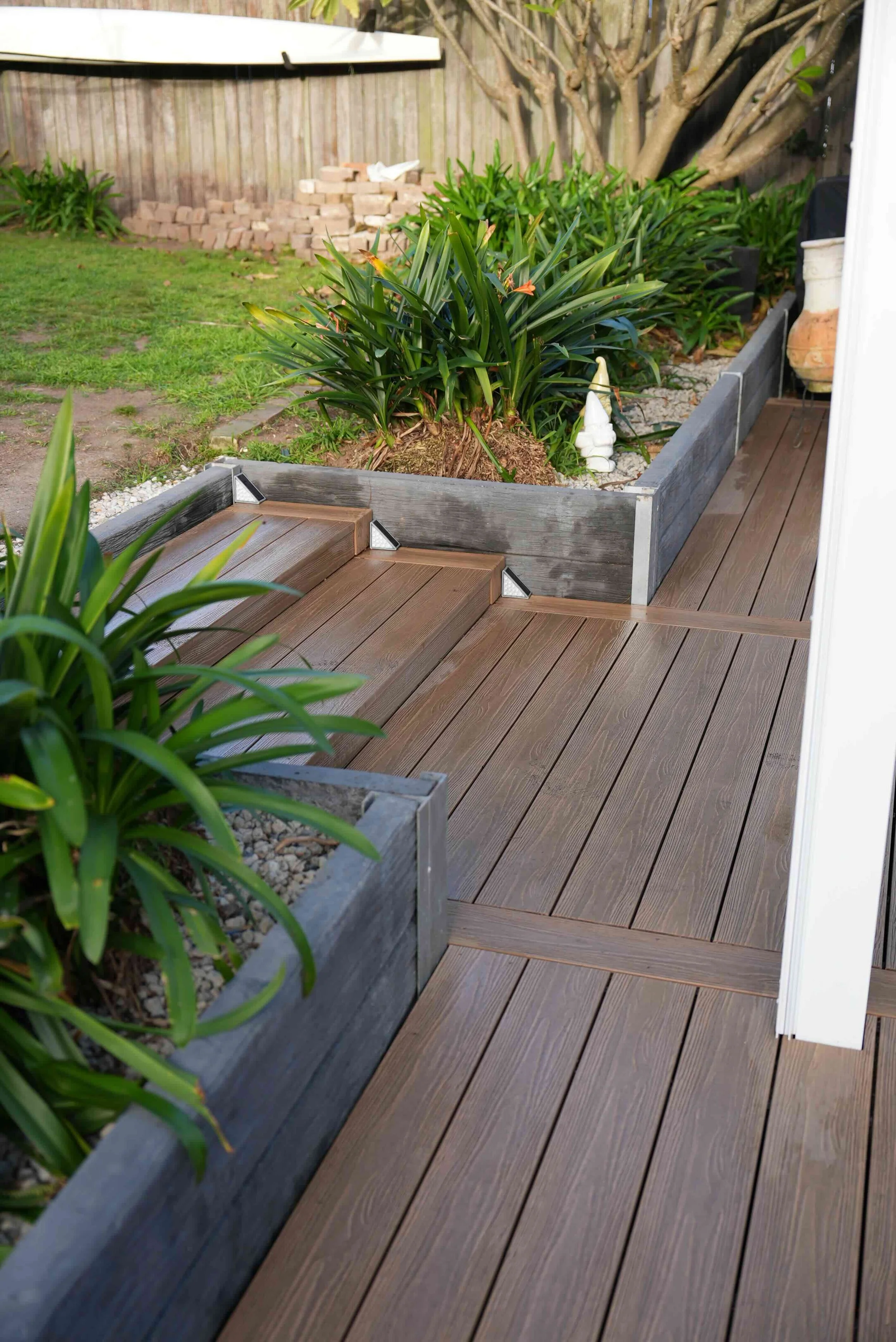 A backyard with a wooden deck, planter box with plants, and a small staircase leading to a grassy area with trees and a stacked pile of bricks in the background.
