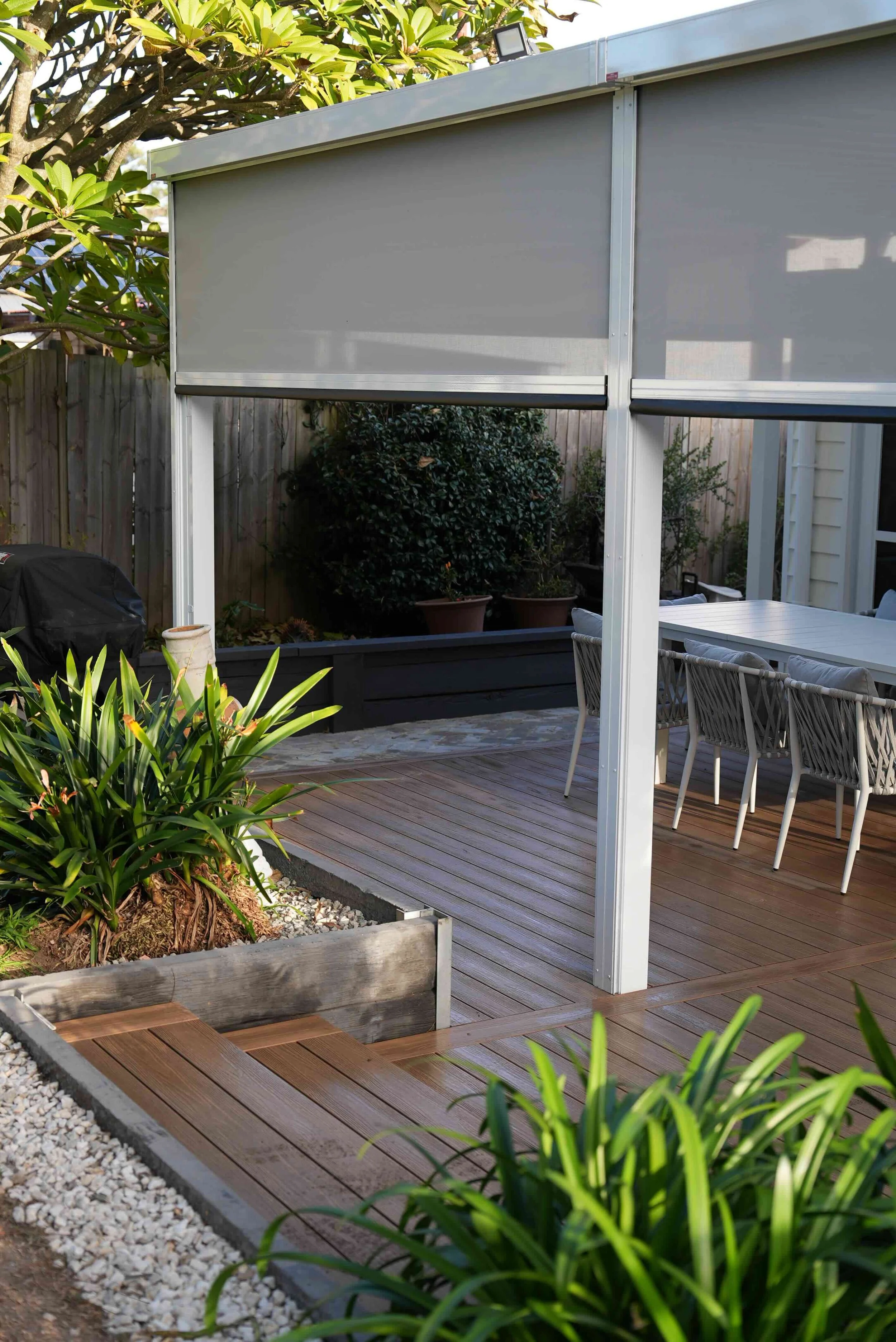 An outdoor patio with a wooden deck, potted plants, garden beds with greenery, a dining table with white chairs, a privacy fence, and a shade cover with roller blinds.